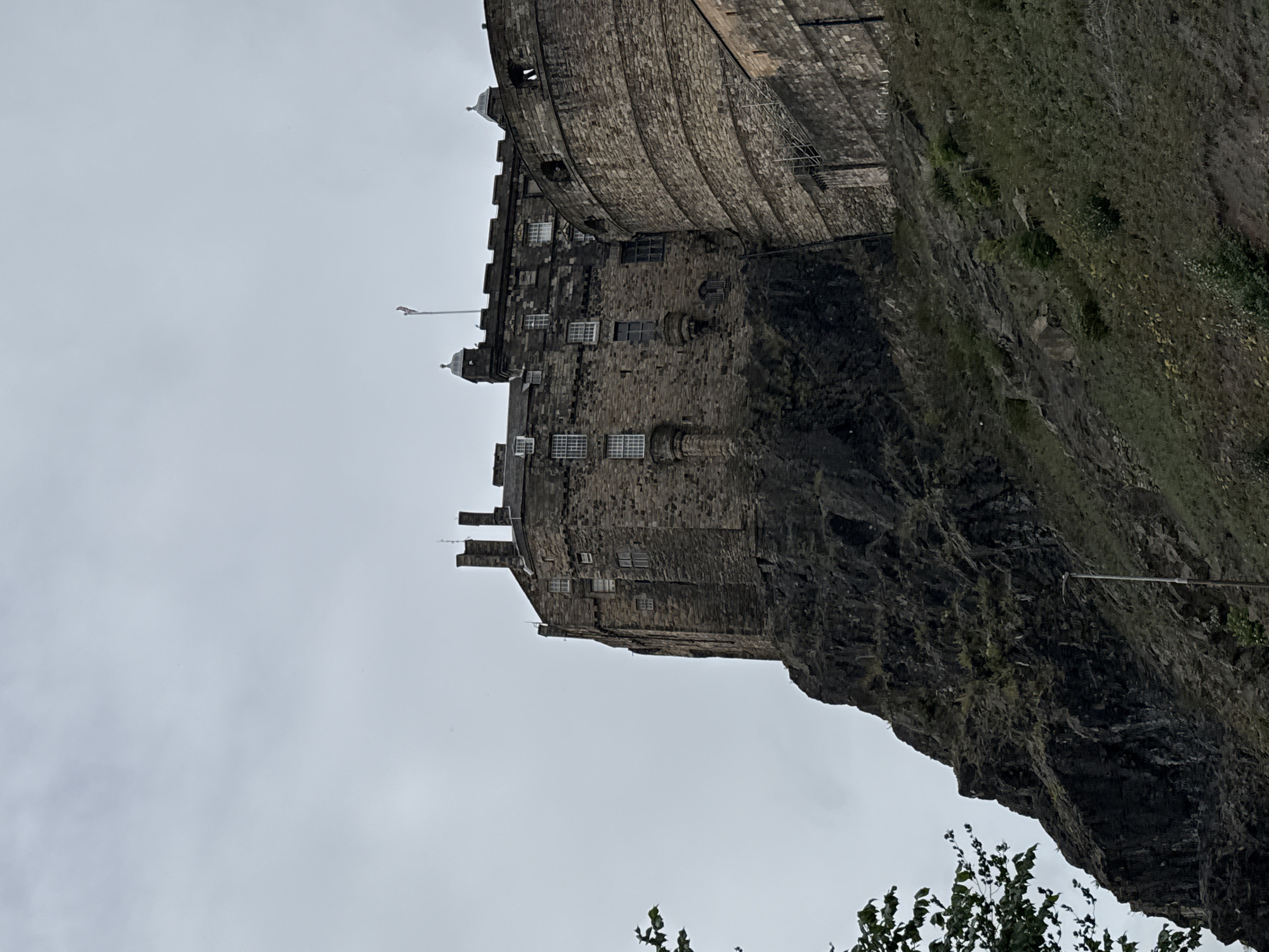 A view of Edinburgh Castle perched atop a steep volcanic rock in the city of Edinburgh, United Kingdom, under a cloudy sky. The castle's stone walls and numerous windows are visible, with a flag flying from a pole on its highest point. The foreground shows a rocky slope covered in sparse vegetation and a lamppost. A tree with green leaves is partially visible on the left side of the image.