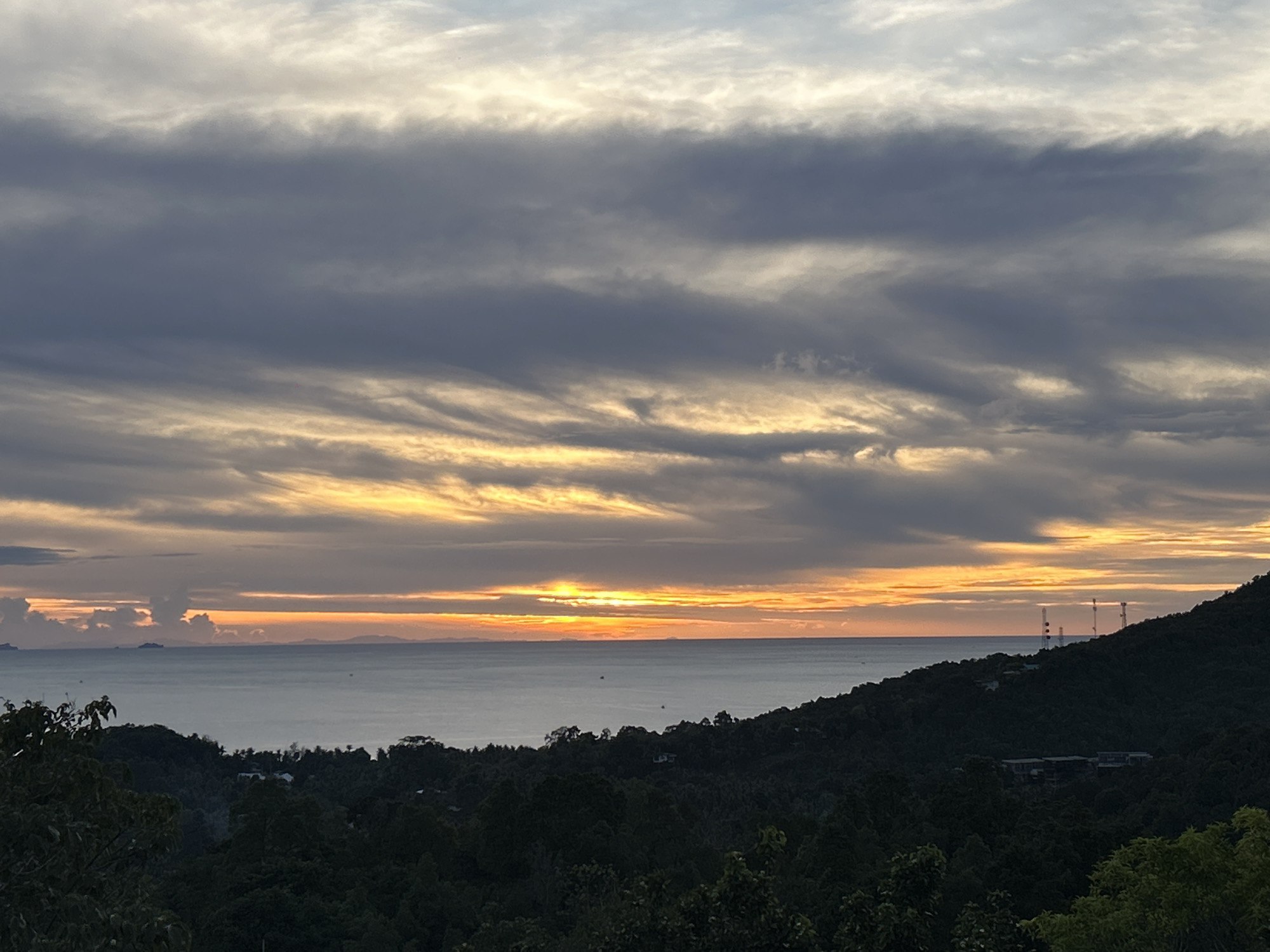 The image shows a sunset over the sea in Baan Sri Thanu, Thailand. The sky is filled with layers of gray and golden clouds. The sun is setting on the horizon, casting an orange glow over the water. Below, a hillside covered in dark green trees is visible. Several cell towers are silhouetted on the right side of the hill.