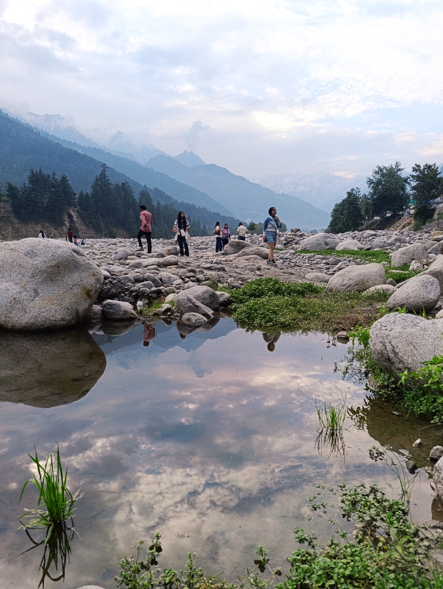 In Vashisht, India, a group of people are gathered on a rocky riverbed with a body of water reflecting the sky and mountains in the background. The scene features a mountainous landscape with trees covering the slopes. Several individuals are scattered across the riverbed, some walking and others standing. The water in the foreground shows a clear reflection of the clouds and the distant mountains. The riverbed is covered with numerous rocks of varying sizes, and patches of green vegetation are visible. The sky is cloudy, suggesting either early morning or late afternoon.