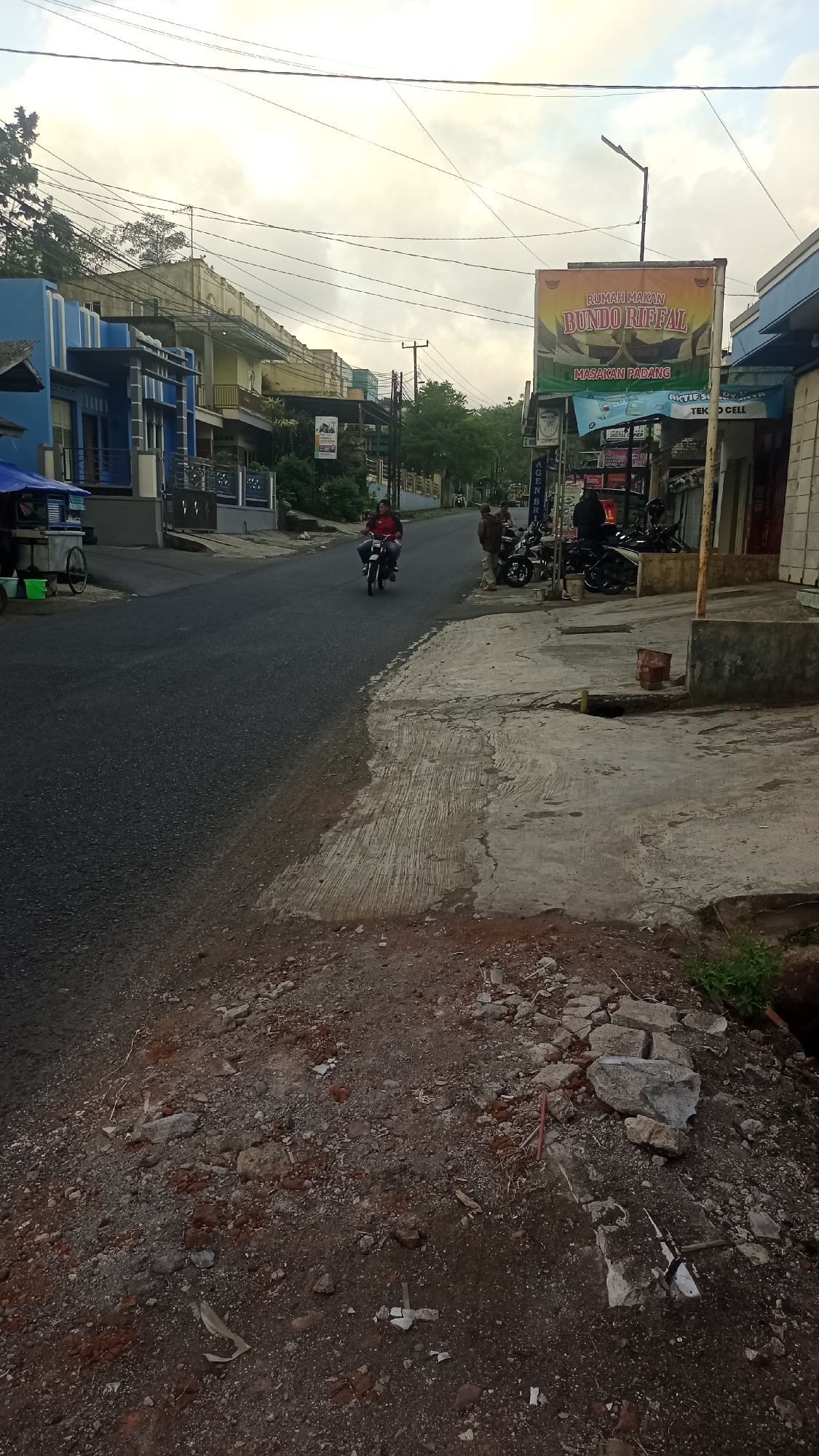 The image captures a street scene in Karyamandala, Indonesia, under an overcast sky. A two-lane asphalt road occupies the foreground and midground, with a concrete sidewalk on the right. The road appears to be newly paved or recently resurfaced, with loose gravel and dirt accumulating on the edges and on the adjacent soil embankment in the foreground.
On the left side of the road, a blue building with white trim and a small blue awning is visible, next to a mobile food cart. Further down the street, a light yellow building with balconies stands. On the right side of the road, a yellow billboard for "RUMAH MAKAN BUNDO RIFAL MASAKAN PADANG" is prominently displayed. Adjacent to the billboard are several shops, one with a sign that reads "TEK O CELL". Motorcycles are parked along the sidewalk in front of these shops.
Two individuals are on the road. One person is riding a motorcycle in the middle of the road, wearing a red shirt. Another person, wearing a brown jacket and a dark shirt, is walking on the sidewalk. Two other individuals are standing on the sidewalk near the parked motorcycles. The environment shows signs of development or repair, with piles of dirt and construction debris present along the roadside. Overhead, a complex network of electrical wires crisscrosses the sky.