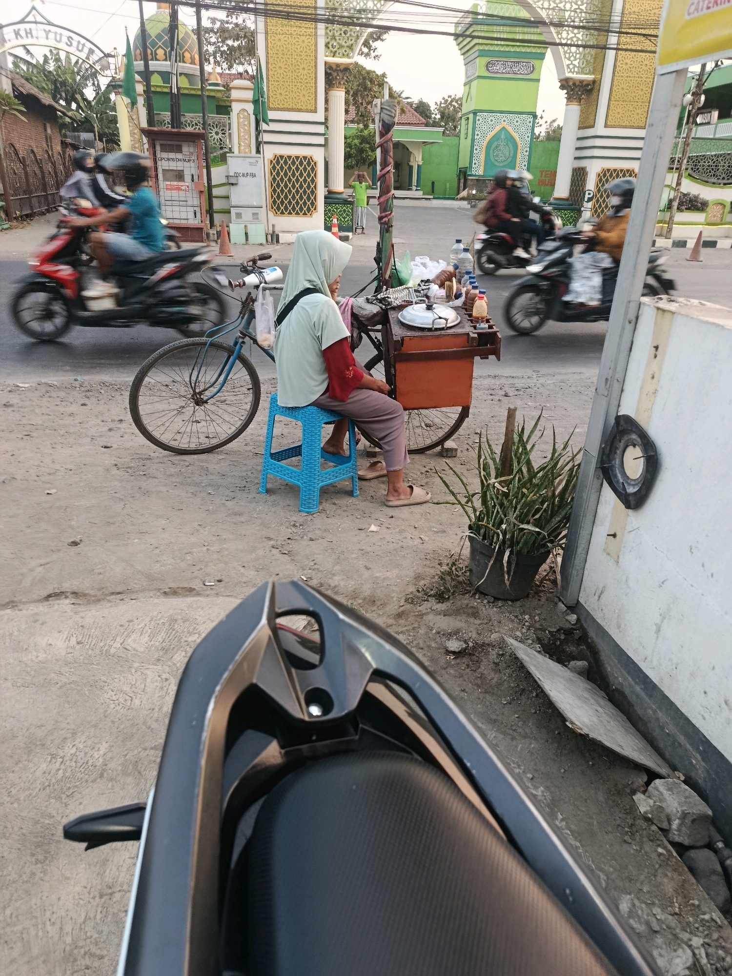 The image depicts a street scene in Kembangarum, Indonesia, with a mosque visible in the background. In the foreground, a person wearing a light green hijab and a red-sleeved shirt sits on a blue plastic stool next to a street food cart. The cart contains various items, including bottles of liquid, a covered pot, and what appears to be food. A blue bicycle is parked behind the cart. Several motorcycles are in motion on the road, with riders wearing helmets. The street is paved with asphalt and concrete. A potted plant with long green leaves is situated near a white wall on the right side of the frame. The overall impression is of a busy, everyday scene in a semi-urban Indonesian setting, likely during the daytime.