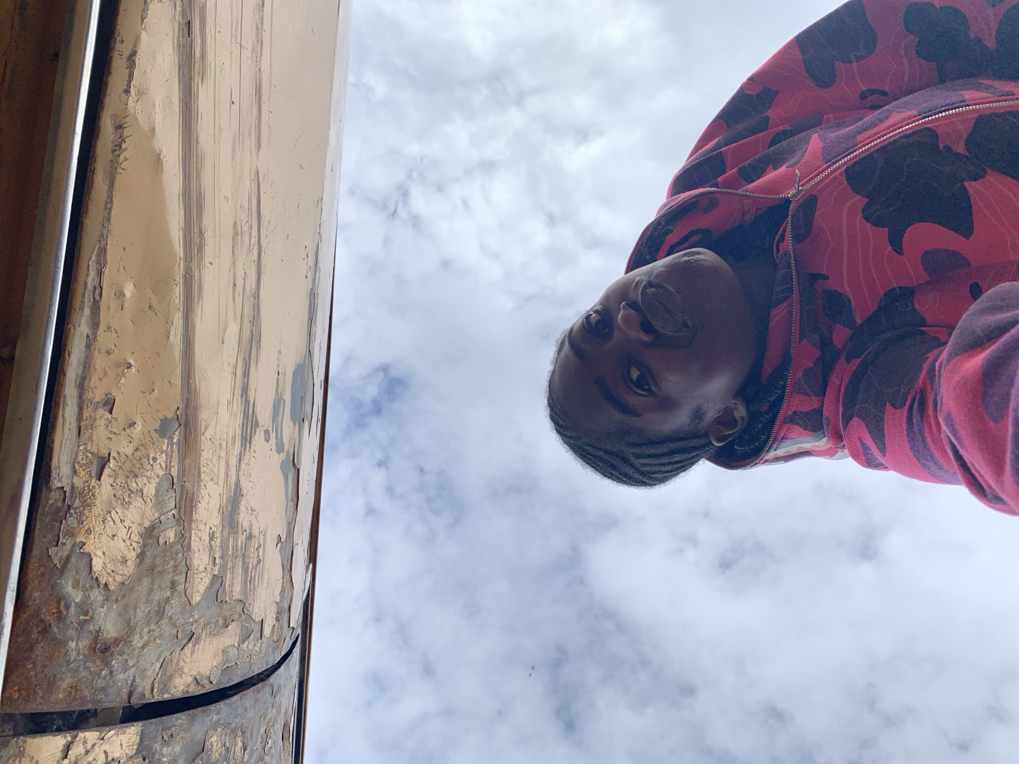 The image, set in Rafin Par, Nigeria, captures a low-angle view looking upward. A dark-skinned individual, appearing to be a young adult, occupies the middle and lower right portions of the frame. The person has braided hair, visible on the left side of their head, and wears a red and pink camouflage-patterned full-zip hoodie, looking directly into the camera with a neutral expression. The upper third of the image is dominated by an overhead structure, possibly a ceiling or overhang, painted a light tan or beige color. This surface exhibits significant deterioration, with large areas of paint peeling and flaking, revealing darker underlayers and extensive surface wear. A distinct vertical seam is visible on the left side of this structure. The background consists of a light blue sky dotted with scattered white clouds.