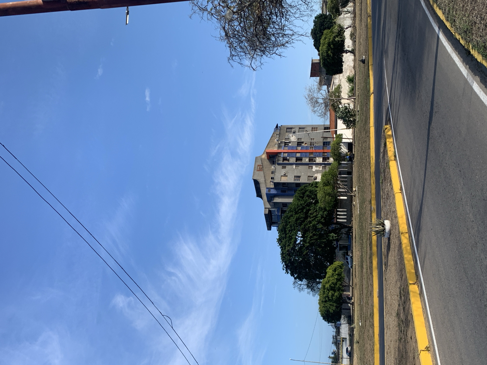 The image depicts a street scene in El Valle del Espíritu Santo, Venezuela, under a bright blue sky with wispy white clouds. In the foreground, a paved road curves to the right, bordered by a yellow and white curb. A patch of dry grass and a small potted succulent mark the median strip.
Dominating the mid-ground is a tall, multi-story building with a gray facade accented by vertical orange and blue stripes. Several trees, including a large, dense green one, are situated in front of and around the building. Behind the building, other structures, including a red-roofed building and a white wall, are visible. The background shows more trees and the distant landscape. A dark pole with a street light is visible on the far right. Power lines stretch across the top of the frame. No people are clearly visible in the scene.