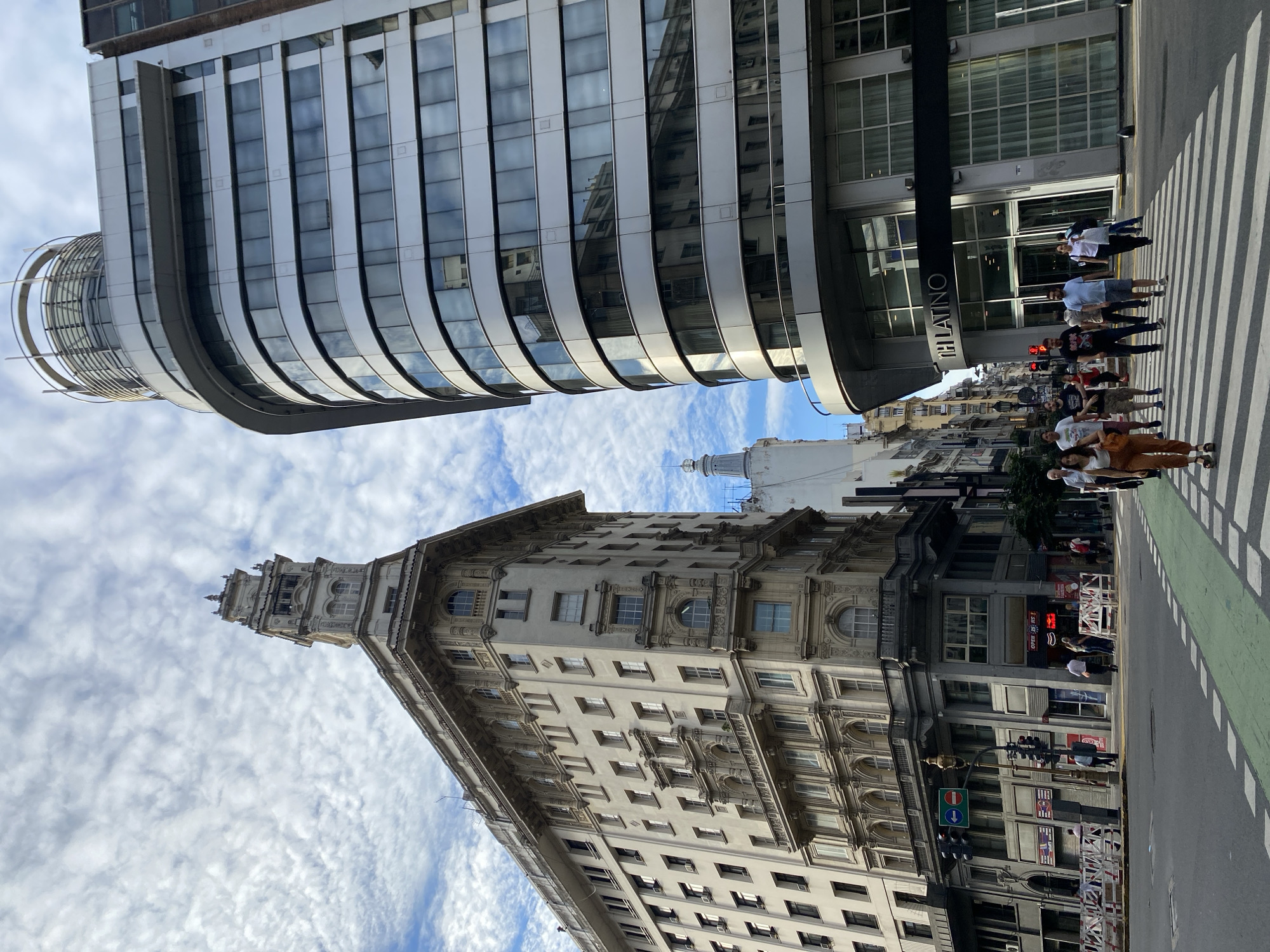 This eye-level shot in Buenos Aires, Argentina, focuses on a street scene framed by two tall buildings. On the left, an older, ornate building rises with a prominent tower. The building on the right exhibits a modern architectural style with a rounded facade. Pedestrians cross a zebra crossing, some observing traffic lights at the intersection. The sky, partly visible between buildings, displays a cloudy pattern. Signage indicates a business named “Latino”.