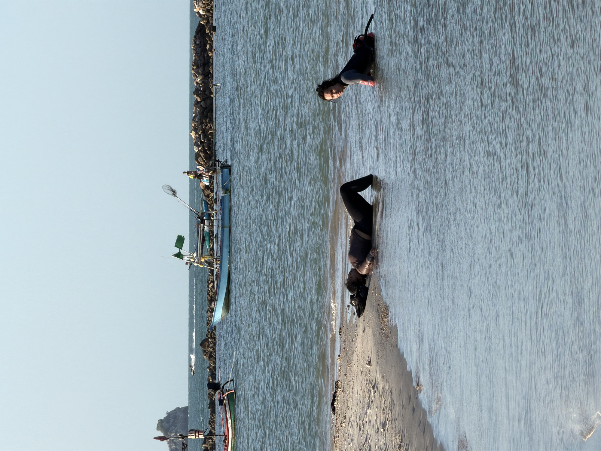 In the foreground of the image, two individuals are present in the shallow, calm water of the ocean. On the left, a person is lying face down on the sandy shore, partially submerged in the water. They are wearing a dark-colored full-body suit. To the right, another person is in a crawling or kneeling position in the water, facing the camera. This individual is also wearing a dark suit with red accents on the arms and appears to be looking towards the shore or the person lying down. The environment is a beach with gentle waves lapping at the shore. In the middle ground, several fishing boats are moored in the water. Some of these boats are small and traditional, while others are larger with fishing equipment visible. In the background, a rocky breakwater stretches into the sea, and beyond that, a jet ski is visible on the water. The sky is clear and blue, indicating a sunny day. The location is identified as the coast of Ko Samui District, Thailand.