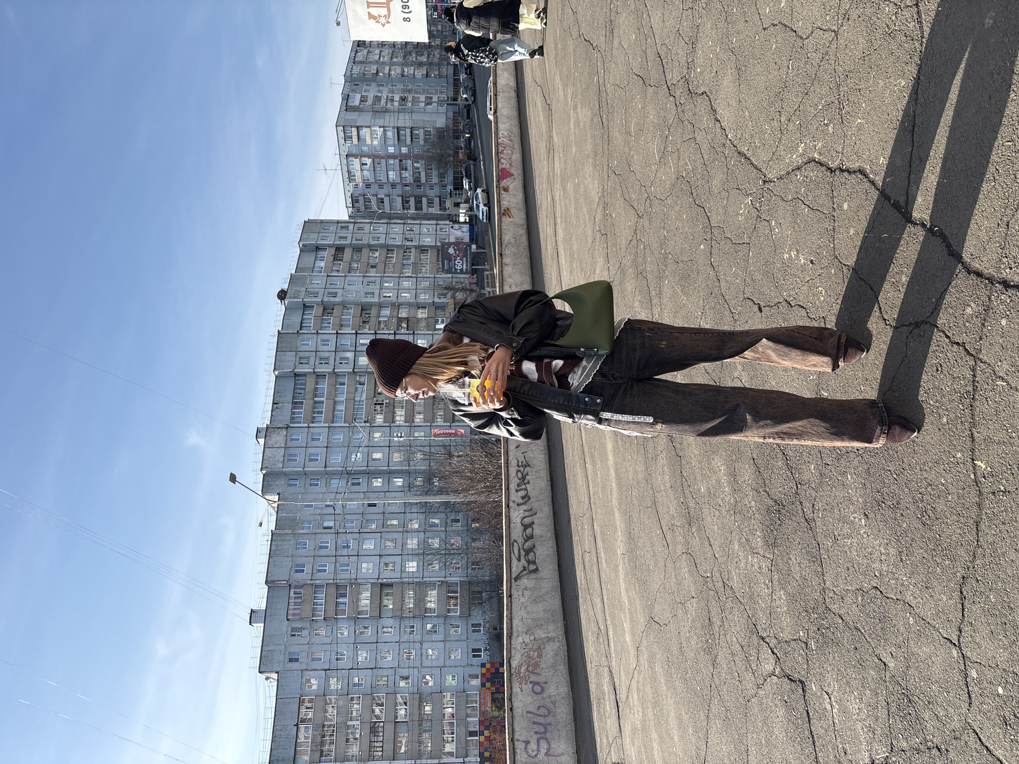 A woman is captured on camera on a sunny day in what appears to be Ust-Ilimsk, Russia, as suggested by the urban landscape in the background. The scene unfolds on a cracked asphalt surface, likely a sidewalk or street. The subject is wearing a brown beanie, a black leather jacket, and brown jeans with brown shoes. She is seen holding a cup, likely filled with an orange beverage, suggesting she is either drinking or about to drink it. A green bag is slung over her shoulder. Behind her, a tall, Soviet-era apartment building rises against the clear blue sky. The building has numerous windows, giving it a grid-like appearance. In the background, there is a street with parked vehicles and a billboard. Graffiti is visible on the wall beside the road. The shadows cast by the person suggest the sun is at an angle.