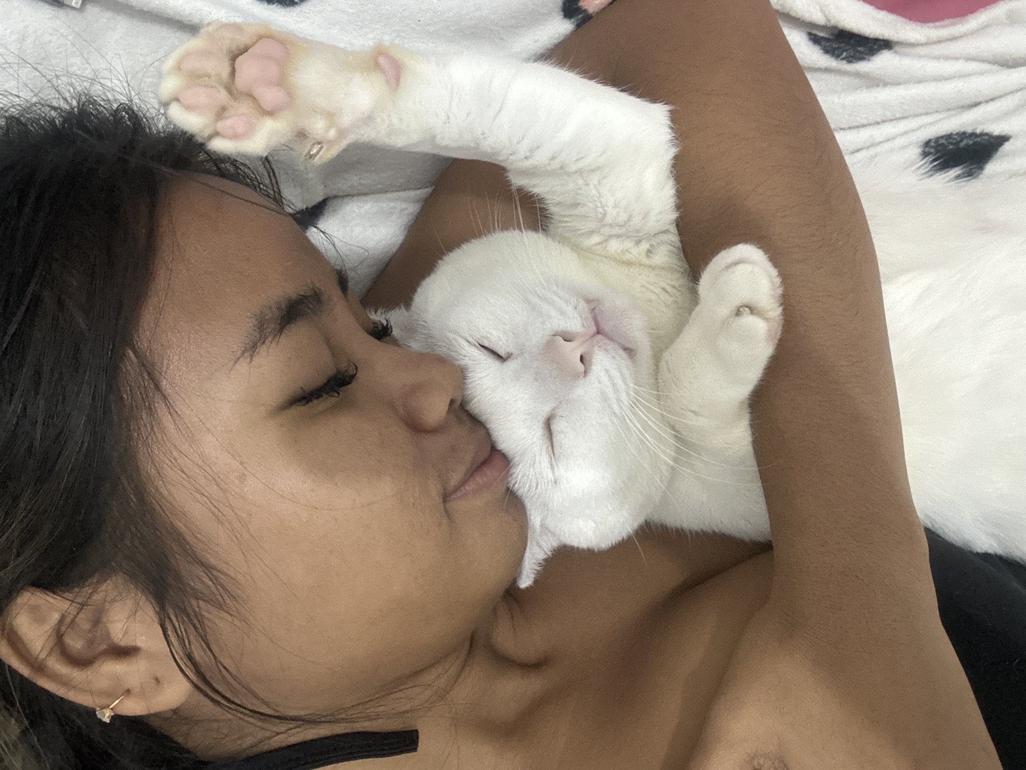 A close-up overhead shot shows a person and a white cat snuggled together on a blanket with a black and white polka dot pattern. The person's face is partially visible, with their eyes closed and head turned to the side. The cat is resting its head on the person's cheek, with its eyes closed and its pink nose and mouth visible. The cat's white paw is draped over the person's arm. The scene appears to be a domestic setting, possibly a bedroom in Sukawati, Indonesia, given the context provided.