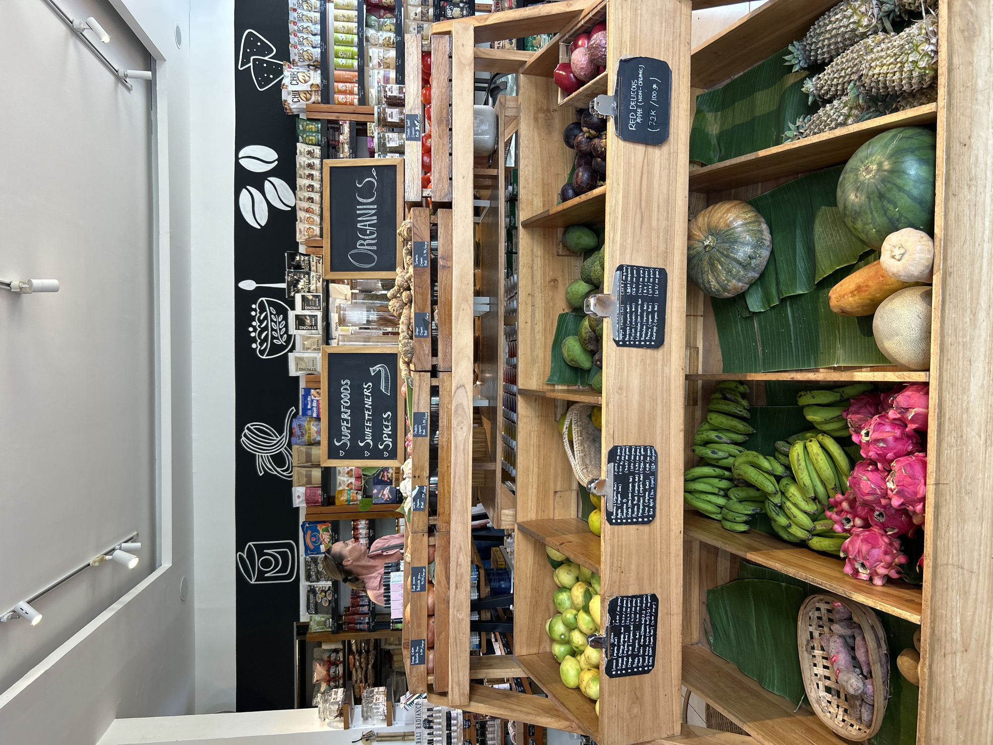 In a brightly lit grocery store in North Kuta, Indonesia, a woman with blonde hair pulled back in a ponytail stands behind a wooden display of fresh produce. She wears a pink collared shirt and appears to be looking to her right. The display, made of tiered wooden crates, holds a variety of fruits and vegetables.
The top crate contains onions and other root vegetables. Below that, a crate is filled with light green citrus fruits, likely limes or lemons, and a basket of similar fruit. Another crate holds avocados and mangosteens, with red apples visible in the far right section.
The second tier of crates showcases more produce. One crate features red tomatoes. Another holds various dried goods and packaged items. A blackboard sign hangs above, reading "ORGANICS" in white chalk.
The bottom tier of crates displays more vibrant fruits and vegetables. One crate contains sweet potatoes in a woven basket, alongside other root vegetables. A crate next to it is packed with bright pink dragon fruits and bunches of green bananas. further to the right, a crate contains a large green watermelon, a papaya, a cantaloupe, and two small round gourds. In the far right, pineapples are arranged neatly.
The background reveals shelves stocked with packaged foods, beverages, and other grocery items. The walls are painted black, with white chalk drawings of food items and text such as "SUPERFOODS," "SWEETENERS," and "SPICES." Track lighting illuminates the scene from the ceiling. The overall impression is that of a well-organized and abundant natural food store.