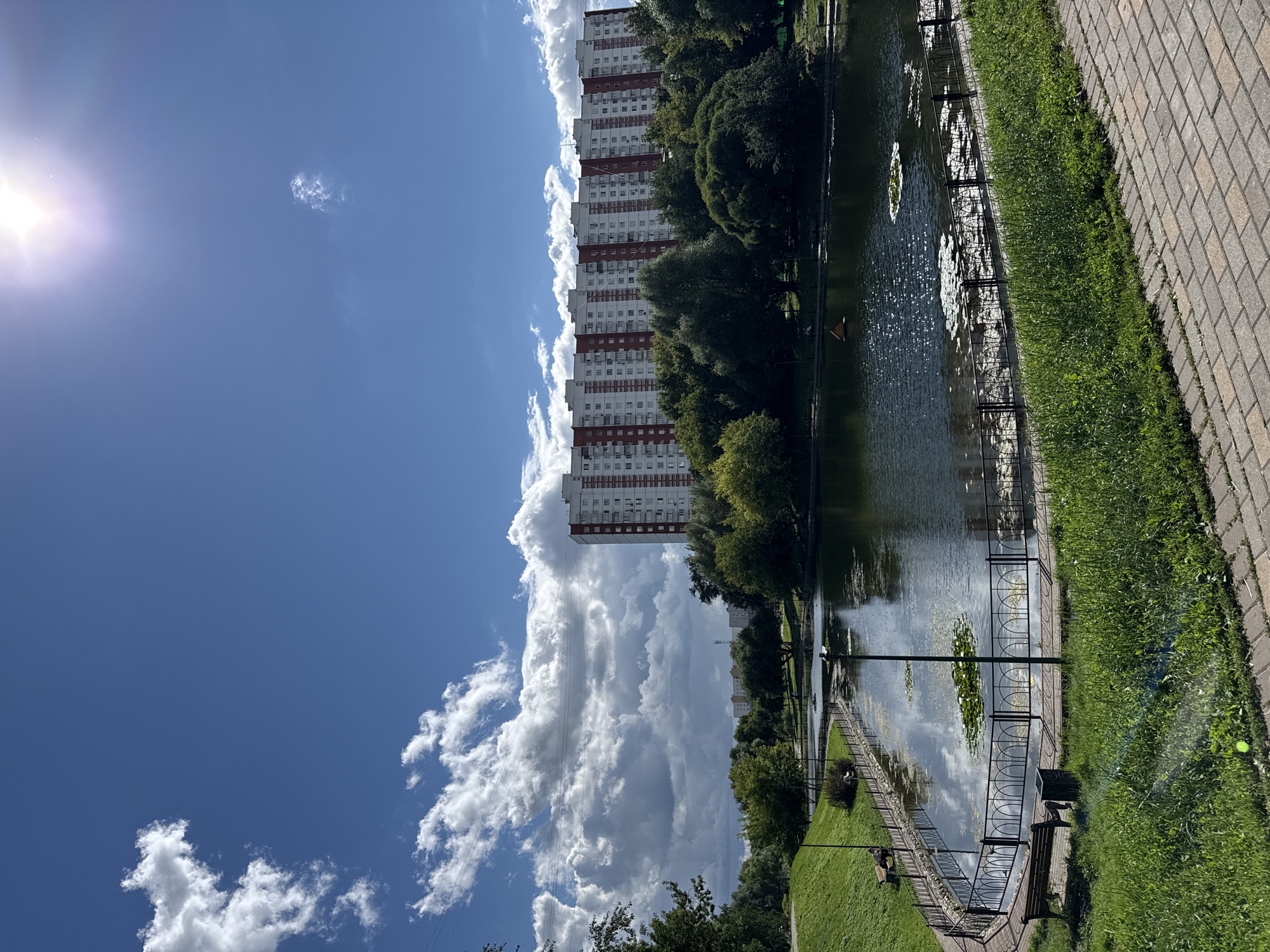 This image captures a sunny day in a city park in Moscow, Russia, featuring a tranquil pond. In the background, a long, multi-story residential building with white and red facades dominates the skyline. Lush green trees and grass surround the pond, with a paved walkway in the foreground. The pond's surface reflects the bright blue sky and scattered white clouds, with a few water lilies visible. Benches are placed along the pond's edge, and a metal fence separates the walkway from the water. The sun is shining brightly, creating a lens flare in the upper right corner of the image. No people are clearly visible in the scene.