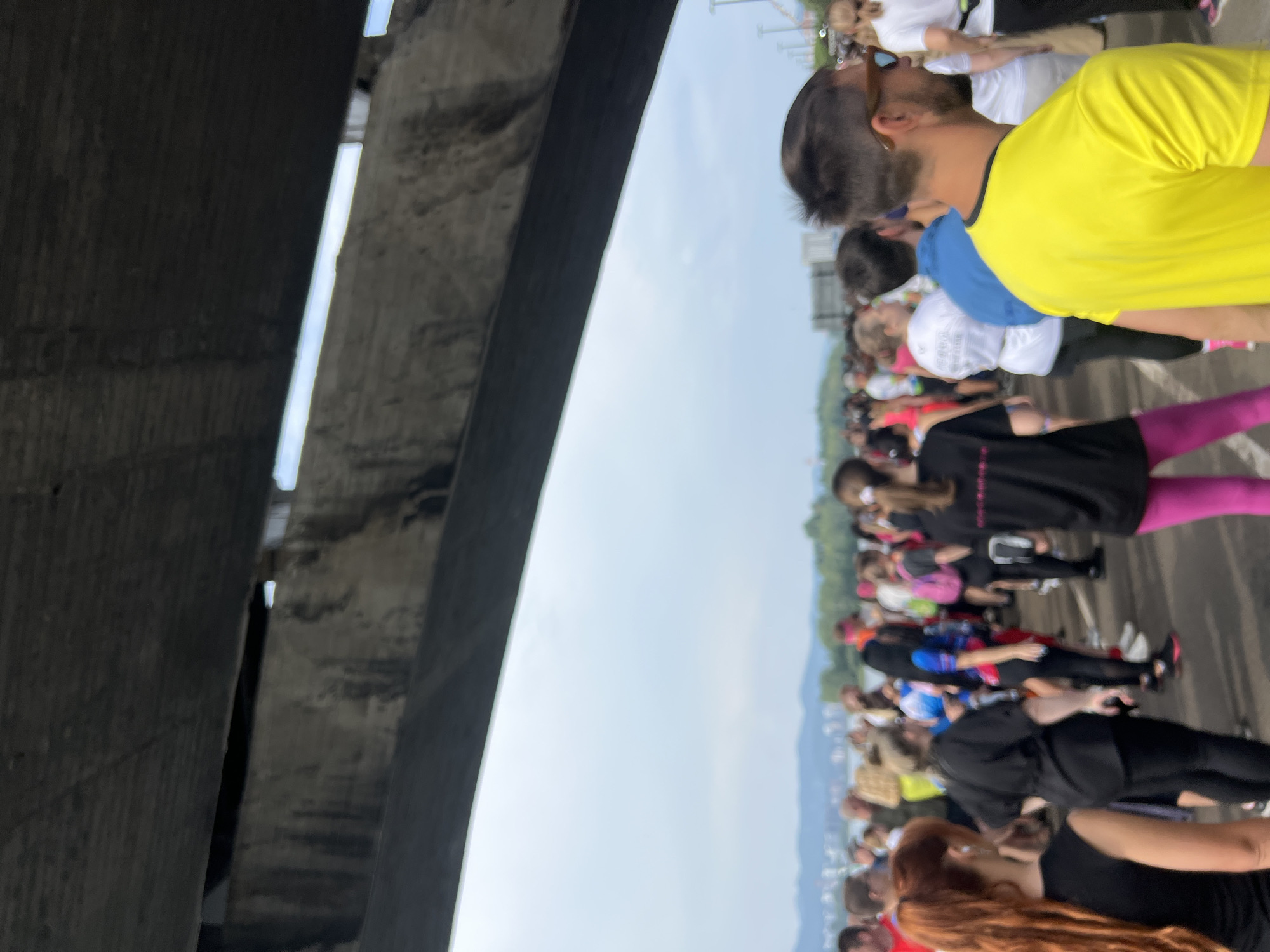 The image captures a gathering of people outdoors, under a large concrete structure, possibly a bridge or overpass. The sky is overcast with a muted light, and in the distance, there is a cityscape with buildings and greenery, suggesting an urban environment.
In the foreground, a man wearing a bright yellow t-shirt and sunglasses is facing to the right. Behind him, other individuals are visible, some wearing t-shirts of various colors, including black, blue, and white. Several people are wearing athletic attire, such as leggings and running shoes, indicating a possible event like a race or a fun run.
The concrete structure overhead is textured and shows signs of wear and tear, with some dark staining. The overall impression is of a casual outdoor event taking place in a city.
There is no specific visual information in the image that allows for the precise identification of the location as Krasnoyarsk, Russia.