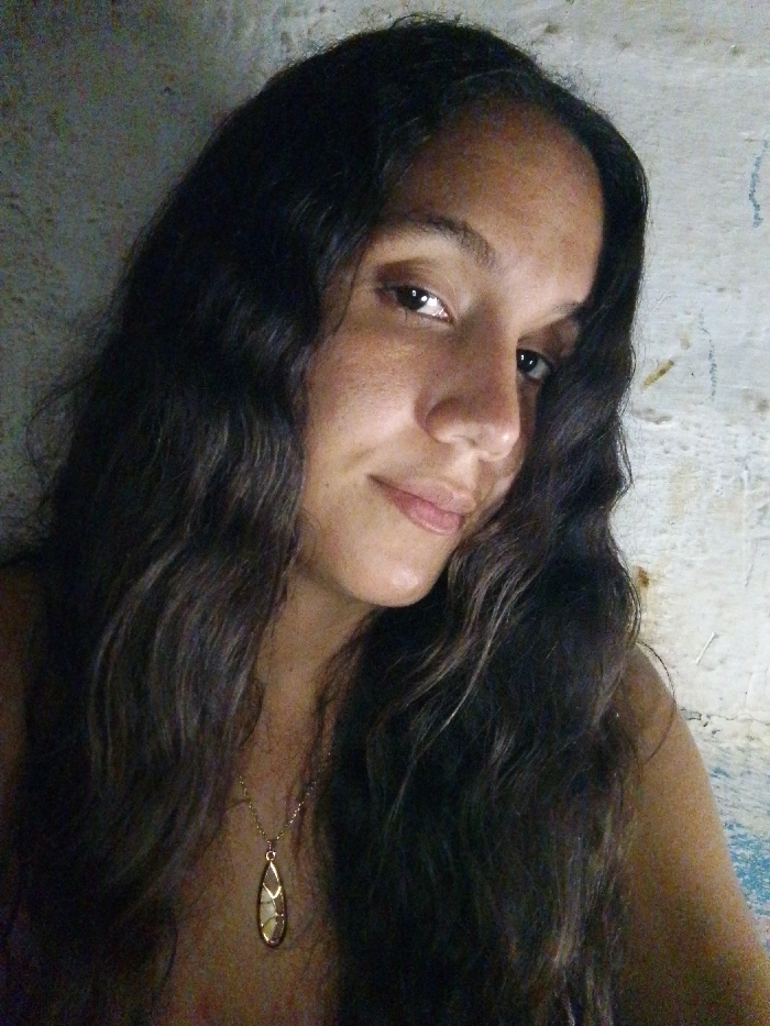 A close-up photograph, taken in Juan Griego, Venezuela, features an adult female subject with long, dark, wavy hair parted on the side and cascading over her shoulders and chest. Her skin is medium-toned with visible freckles across her nose and cheeks. She gazes directly at the viewer with a subtle smile and her head slightly tilted. She wears a thin golden necklace with an elongated, tear-drop shaped pendant featuring a gold frame around a pale, possibly cream-colored, central element. The necklace rests on her upper chest. The background is an unevenly lit, textured wall, appearing dark grey on the left side of the frame and off-white with faint vertical blue discolorations on the right. The primary light source illuminates the subject's face from the front.