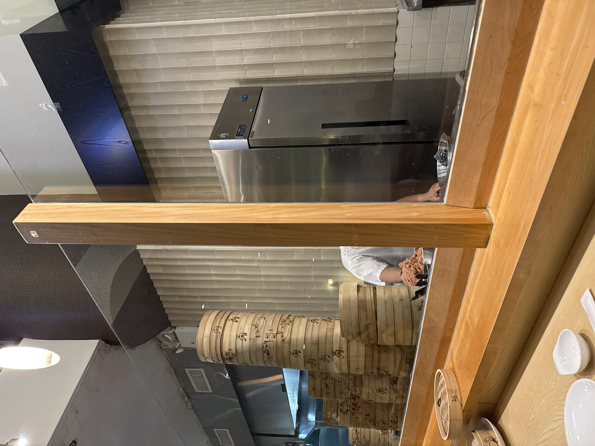 The image depicts a restaurant scene, likely located in New York City, United States of America. Through a glass barrier, a cook in a white coat is preparing food in a metallic bowl. Stacks of bamboo steamers with various designs line the lower portion of the image, visible through the glass. Stainless steel equipment, possibly a refrigerator, is also seen in the background. Wooden frames surround the glass, adding depth to the composition. On the right side of the frame are small white bowls and other dining accessories on a wooden counter.