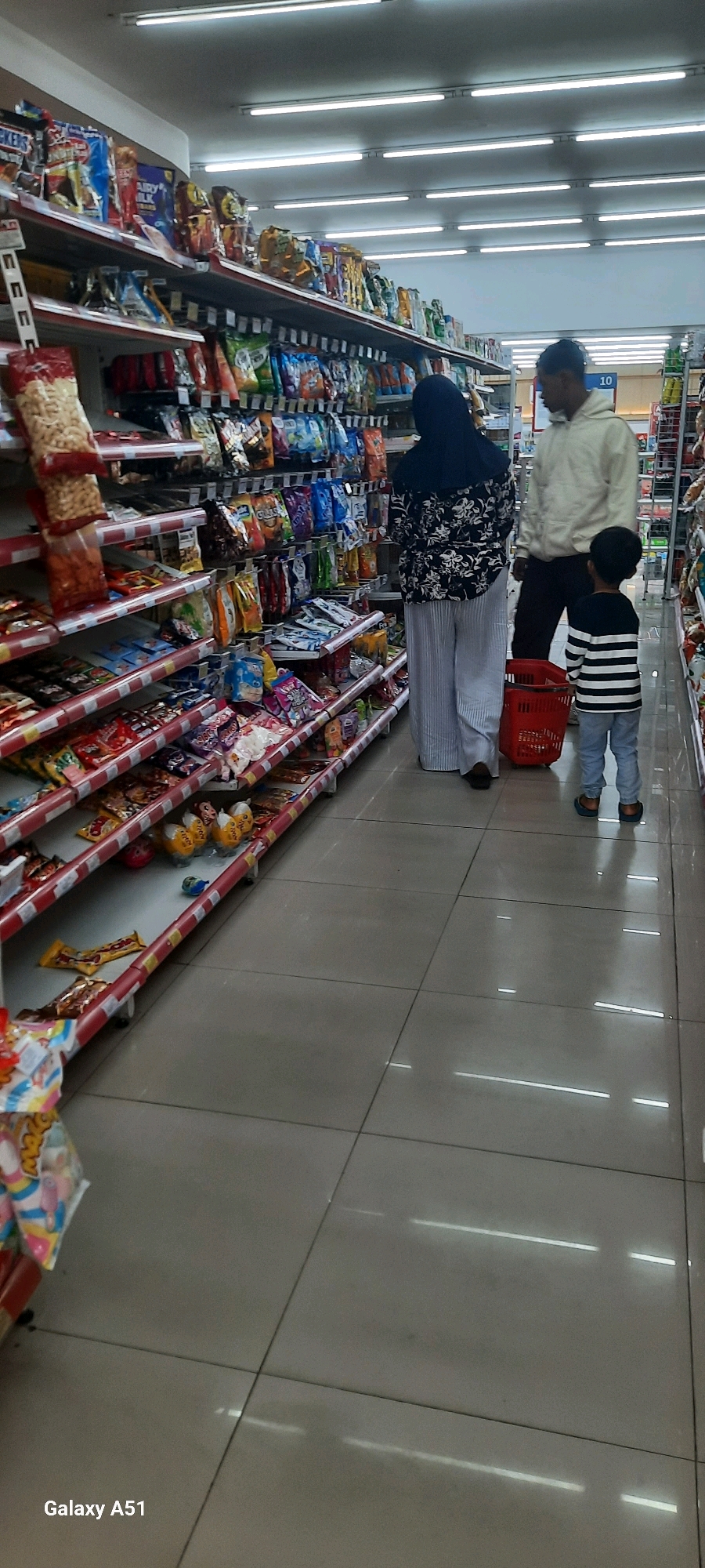 This image depicts a family, likely a mother, father, and child, browsing in a brightly lit convenience store. The shelves are stocked with various packaged snacks and food items. The mother, wearing a dark hijab and a patterned top with wide-legged striped pants, stands looking at the shelves. The father, in a light-colored hooded sweatshirt, stands beside her. A young child, wearing a striped long-sleeved shirt and light-colored pants, stands near a red shopping basket. The flooring is tiled and reflective, with overhead fluorescent lights creating long streaks of light. The overall impression is a typical scene of a family shopping for groceries. The store appears to be a modern retail establishment, possibly a minimarket, common in urban and semi-urban areas.