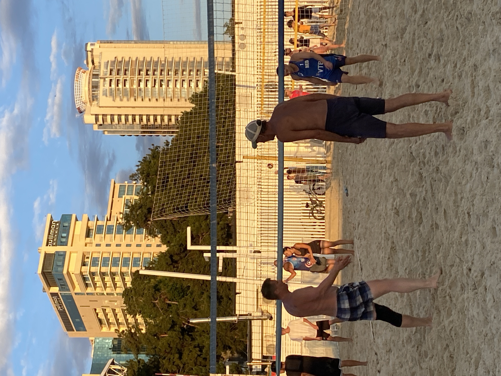 The image depicts a beach volleyball game on a sandy court, set against the backdrop of Sochi, Russia. Two men are in the foreground, facing away from the camera. The man on the left is wearing checkered shorts and a black knee sleeve, while the man on the right is wearing dark blue shorts and a grey baseball cap. Behind them, a volleyball net divides the court, and several other players are visible in the background, some engaged in the game.
The scene is illuminated by the warm glow of the setting sun, casting long shadows on the sand. The environment features tall, modern buildings, including the Pullman Hotel, characteristic of Sochi's urban landscape. Lush green trees surround the beach area, adding a touch of nature to the scene. The sky above is a clear blue with scattered white clouds, suggesting pleasant weather. In the middle ground, a white fence and a bicycle are also visible, further contributing to the beachside atmosphere. The overall impression is one of a leisurely outdoor activity taking place in a resort city.