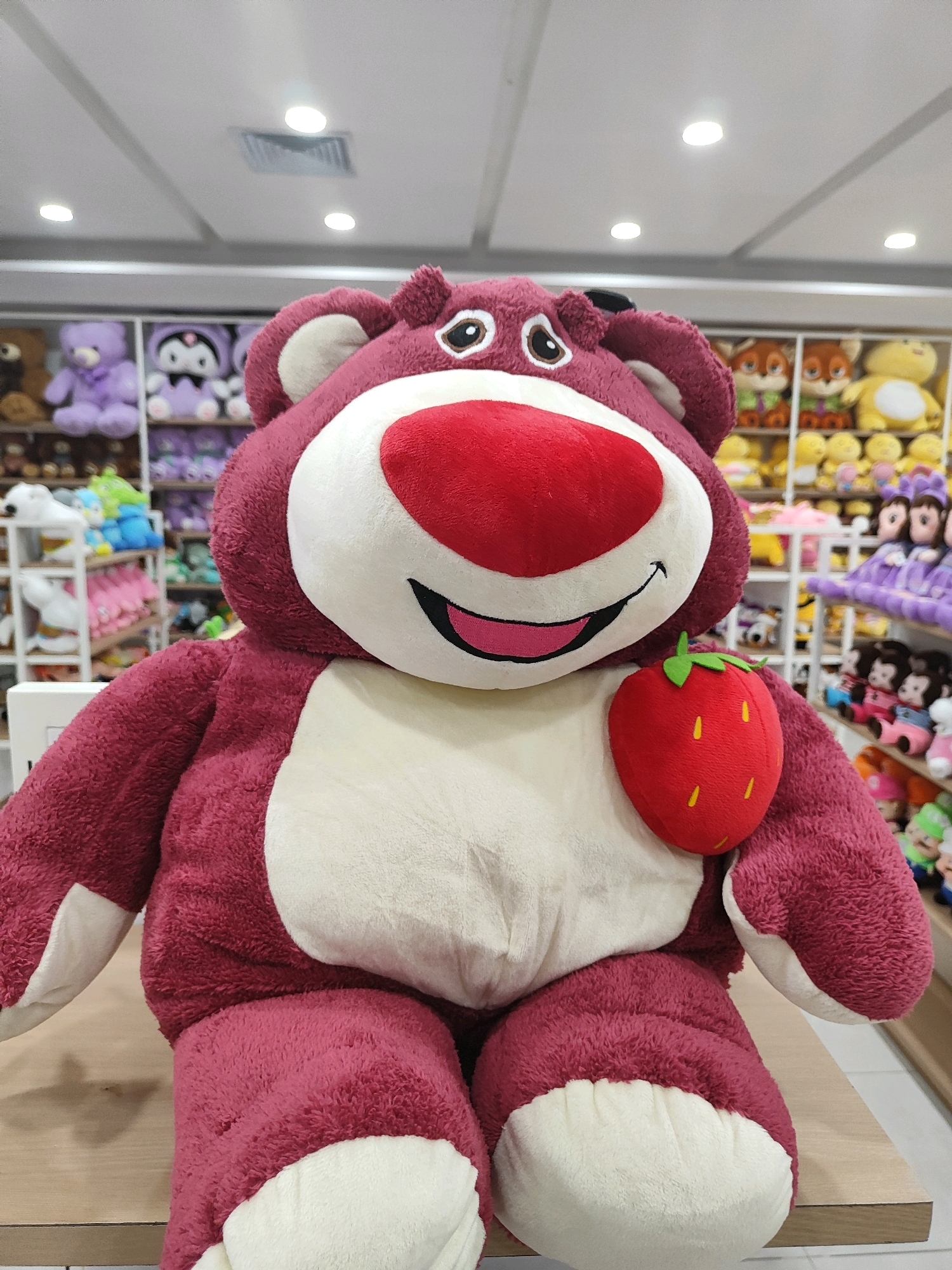 A large, plush Lotso bear, a character from Toy Story, is the central focus of the image. The bear is a dark pink color with a cream-colored belly, snout, and inner ears. It has a prominent red nose and black eyes with white pupils. A small, red strawberry with green leaves is attached to its chest. The bear is seated on a wooden surface, likely a shelf or table, in what appears to be a store selling stuffed animals. In the background, rows of other stuffed animals are visible on shelves, including teddy bears, cartoon characters, and other plush toys. The lighting in the store is bright and comes from overhead fluorescent lights. The overall scene suggests a retail environment filled with a variety of toys. The location is identified as Sukowiryo, Indonesia.