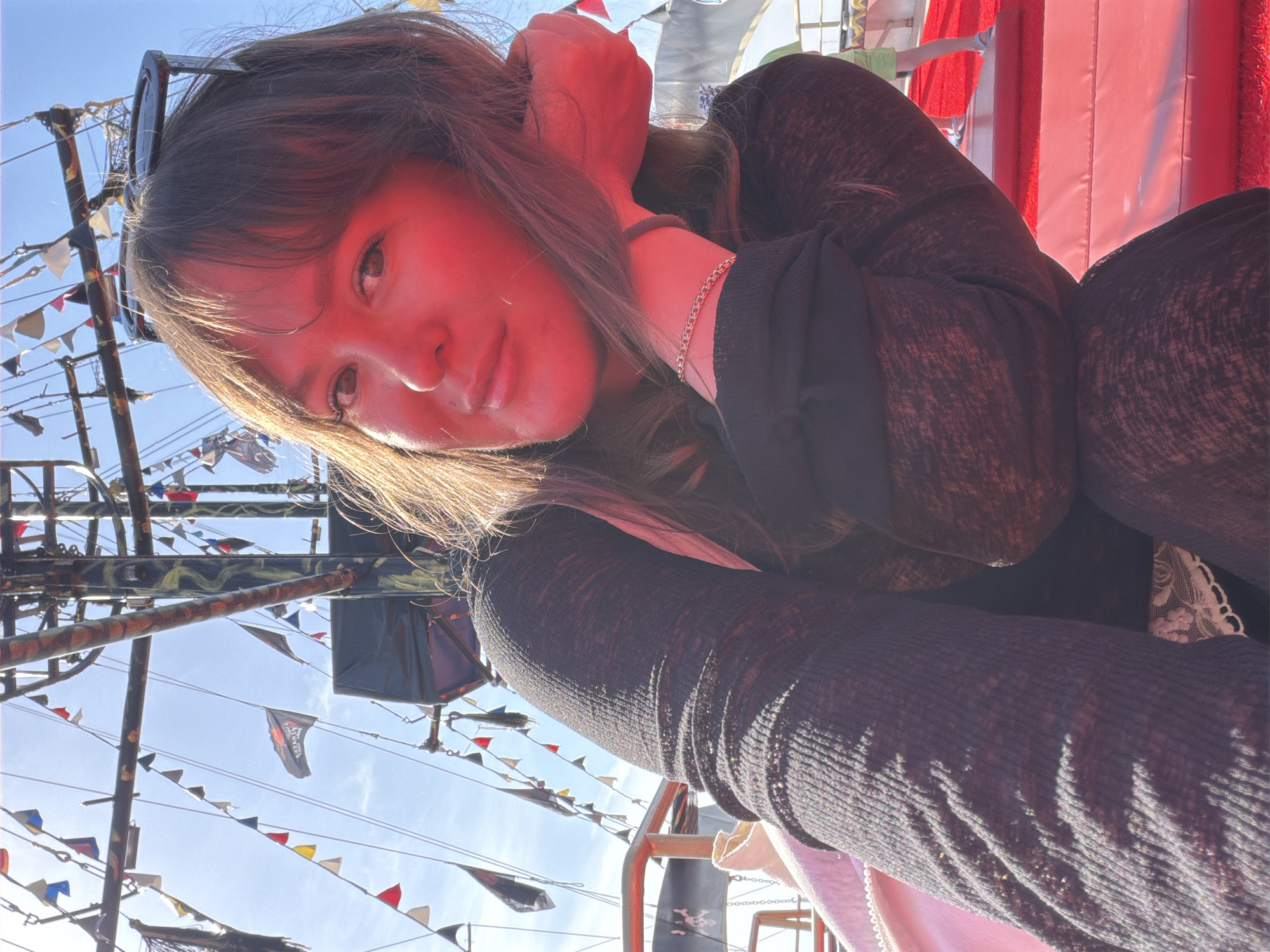 This is a brightly lit selfie taken by a young woman on what appears to be a pirate-themed tour boat in Alanya, Turkey. The image is taken from a low angle, looking upwards at the subject and the sky.
The main subject is a woman with long, dark brown hair, partially covering her forehead with bangs. She has sunglasses perched on top of her head. She is looking directly at the camera with a subtle smile. Her face is illuminated by strong sunlight, casting a reddish hue on her skin. She wears a dark, possibly ribbed or textured, long-sleeved top and a delicate chain bracelet on her left wrist.
The background reveals a clear, bright blue sky. Numerous ropes, masts, and rigging of a boat are visible, festooned with strings of colorful pennants (small triangular flags) in red, yellow, blue, and white. Several larger black flags are also present, some clearly displaying the word "PIRATES" in white text and featuring a skull and crossbones emblem, reinforcing the pirate ship theme. To the right, a large red inflatable structure, possibly part of a slide or bouncy castle, is visible, suggesting entertainment facilities on board.
The time of day is sunny, indicated by the clear sky and bright highlights on the woman's hair and the boat's rigging. The overall mood appears relaxed and cheerful, consistent with a leisure activity like a boat tour.