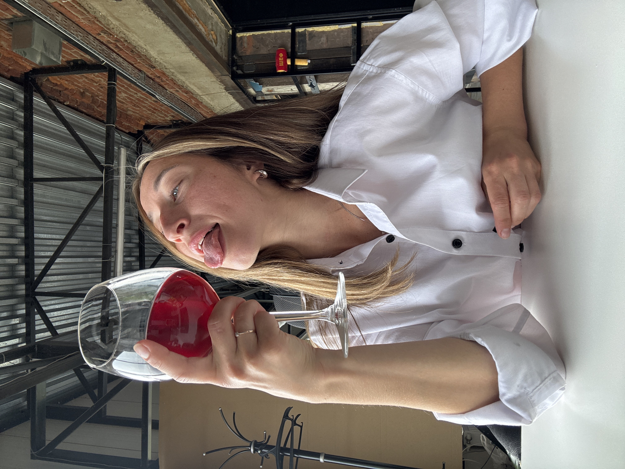 A young woman with long, blonde-streaked brown hair sits at a white table in an urban setting. She is wearing a white collared shirt, unbuttoned at the top. In her right hand, she holds a large wine glass filled with a red liquid, tilting it upwards. Her head is tilted back, and her tongue is sticking out towards the glass. Behind her, a brick wall is visible, along with exposed metal beams and corrugated metal roofing, suggesting an industrial or loft-style interior. A coat rack stands to the left, and a red object resembling a hammer with Cyrillic lettering is mounted on the wall in the background. The overall atmosphere is casual and playful.