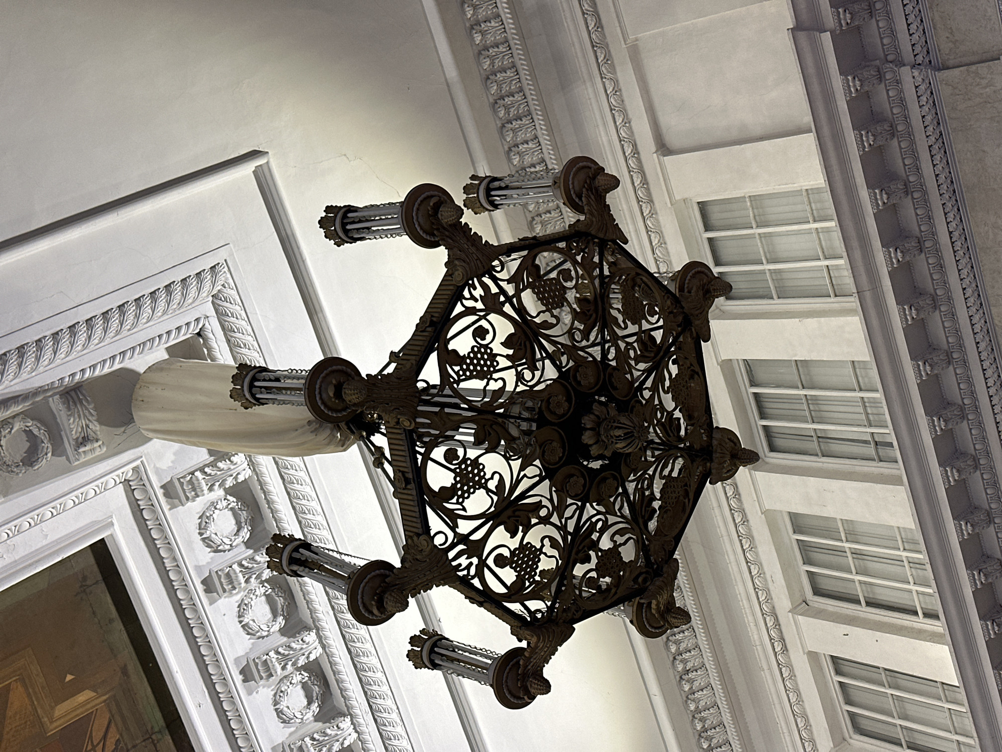 A low-angle shot captures an ornate chandelier suspended from the ceiling. The chandelier has an elaborate metal framework with a design featuring grape vines and flowers. Cylindrical lights extend from the chandelier's frame. The ceiling around the chandelier is decorated with intricate molding and architectural details, including a series of framed rectangular windows. This location is likely a historical or monumental building in Saint Petersburg, Russia.