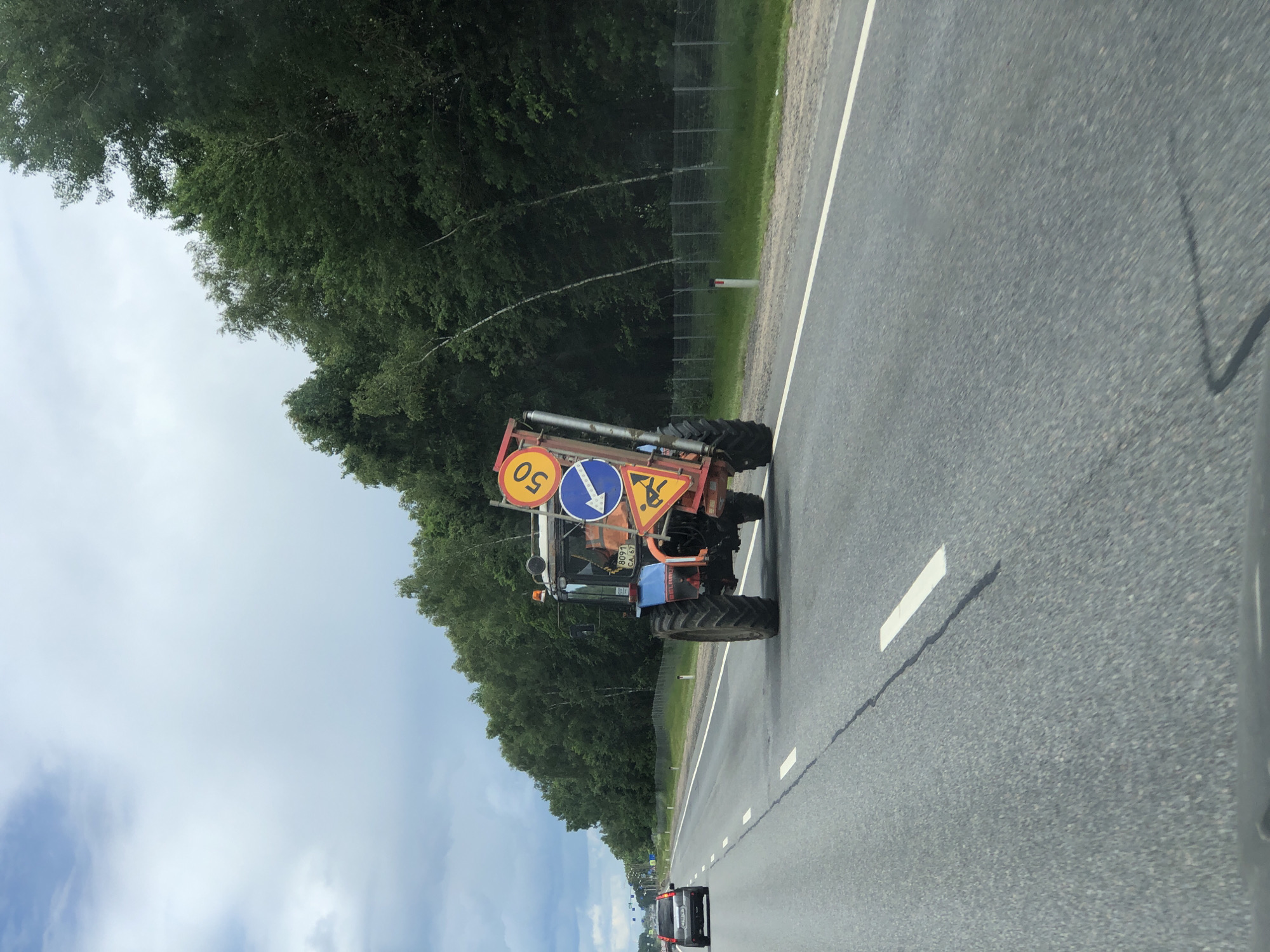 A blue tractor with construction signs is driving on a highway. One sign indicates a speed limit of 50 kilometers per hour. Another sign shows a blue arrow pointing to the right, and a third sign depicts a person working. The tractor is carrying a red trailer attached to its rear. The tractor has a license plate with the number "8091 CA 67". The scene takes place on a cloudy day on a paved highway. Trees line the side of the road, and a wire fence separates the road from the trees. A silver car is visible driving ahead of the tractor. The location is Kablukovo, Russia.