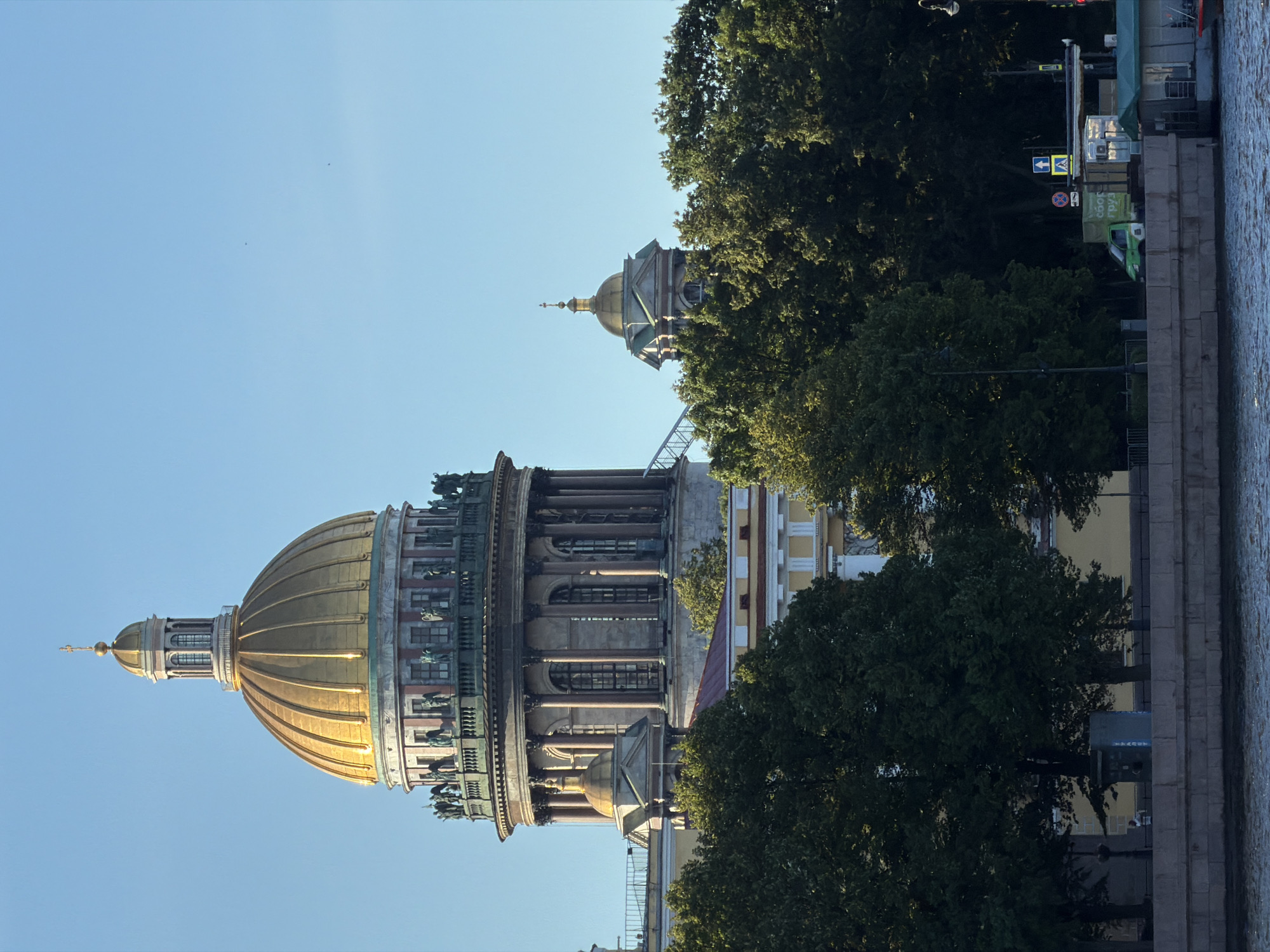 The image shows Saint Isaac's Cathedral in Saint Petersburg, Russia, as viewed from across a body of water, likely the Neva River. The large, golden dome of the cathedral dominates the upper left of the frame, with a smaller golden dome of a secondary structure visible to its right, peeking through lush green trees. The sky is a clear, bright blue, suggesting fair weather. In the foreground, a stone embankment lines the water, with trees obscuring much of the background. A few small boats and a green truck are visible near the water's edge, along with some street signs and a kiosk. No people are prominently featured or engaged in significant actions. The scene depicts a classic architectural landmark bathed in sunlight.