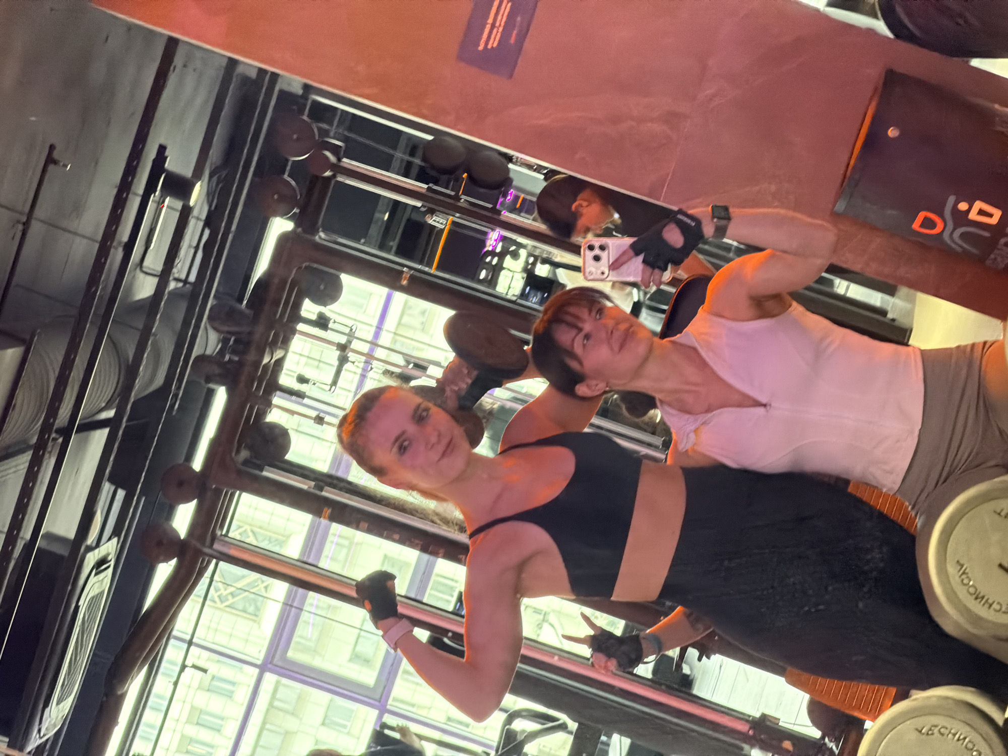The image depicts two women posing in a modern gym in Perm, Russia, during the daytime. The camera's orientation is tilted, presenting the scene diagonally.
In the foreground, a woman with light brown hair, pulled back, is wearing a black sports bra and black patterned leggings. She has black fingerless gloves on both hands, with a white wristband on her left arm. She holds a dumbbell in her right hand, flexing her arm, and looks towards the camera with a faint smile.
Behind her and to the right, another woman with darker hair and bangs is seen. She wears a white top, light brown shorts, black fingerless gloves, and a dark smartwatch on her left wrist. She is holding a white smartphone, appearing to take a mirror selfie or video, with her reflection visible in the large mirror behind her.
The gym features an industrial design with exposed metal frameworks, pipes, and ductwork. Large windows in the background reveal the bright daytime sky and modern high-rise buildings, suggesting an urban setting. To the right of the women, a rack of dumbbells is visible, some imprinted with the brand name "TECHNOGYM" (partially inverted). A black sign or poster is visible in the background with partially legible white and orange letters that appear to spell "D.O." and "U". Another sign, partially visible in the top right, shows Russian text: "ПРИВЕТСТВУЕМ ВАШУ" and "БЕЗОПАСНОСТИ" (meaning "Welcome your" and "safety" respectively). The overall atmosphere suggests an active and confident workout session.
