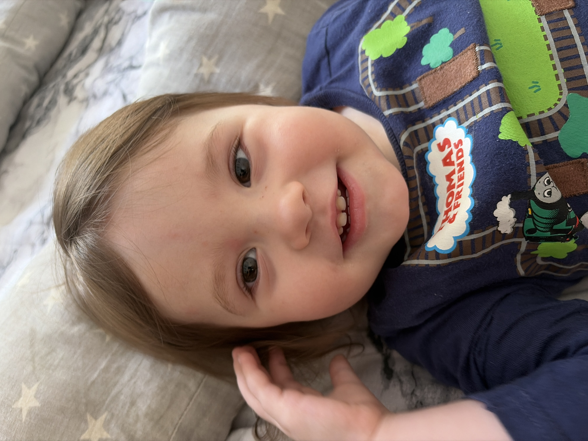 A toddler lies on a pillow with a star pattern, wearing a blue shirt with a "Thomas & Friends" design featuring railroad tracks, trees, and the titular character. The child is smiling, showing upper teeth. The setting appears to be indoors, possibly a home. There is no information about Chaikovsky, Russia in the scene.