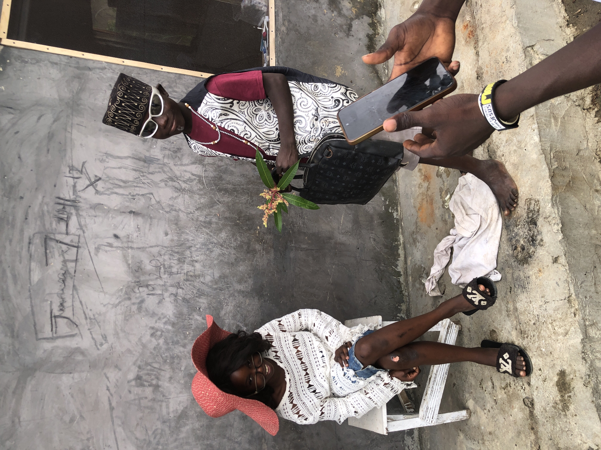 In Ado, Nigeria, two children are posing for a photograph against a textured, grey concrete wall. The child on the left, a girl, is seated on a white stool, wearing a large, floppy pink hat, glasses, a white crocheted top, and denim shorts. She is looking at the camera and smiling. The child on the right, a boy, stands and wears a dark cap with ornate gold patterns, white sunglasses, a maroon t-shirt, and a patterned vest. He is holding a bouquet of flowers in his left hand and a black handbag in his right. In the foreground, two sets of hands are visible, one holding a smartphone, suggesting someone is taking a picture of the two children. A white cloth lies on the ground near the girl's feet. The wall behind them has some faint, scrawled markings.