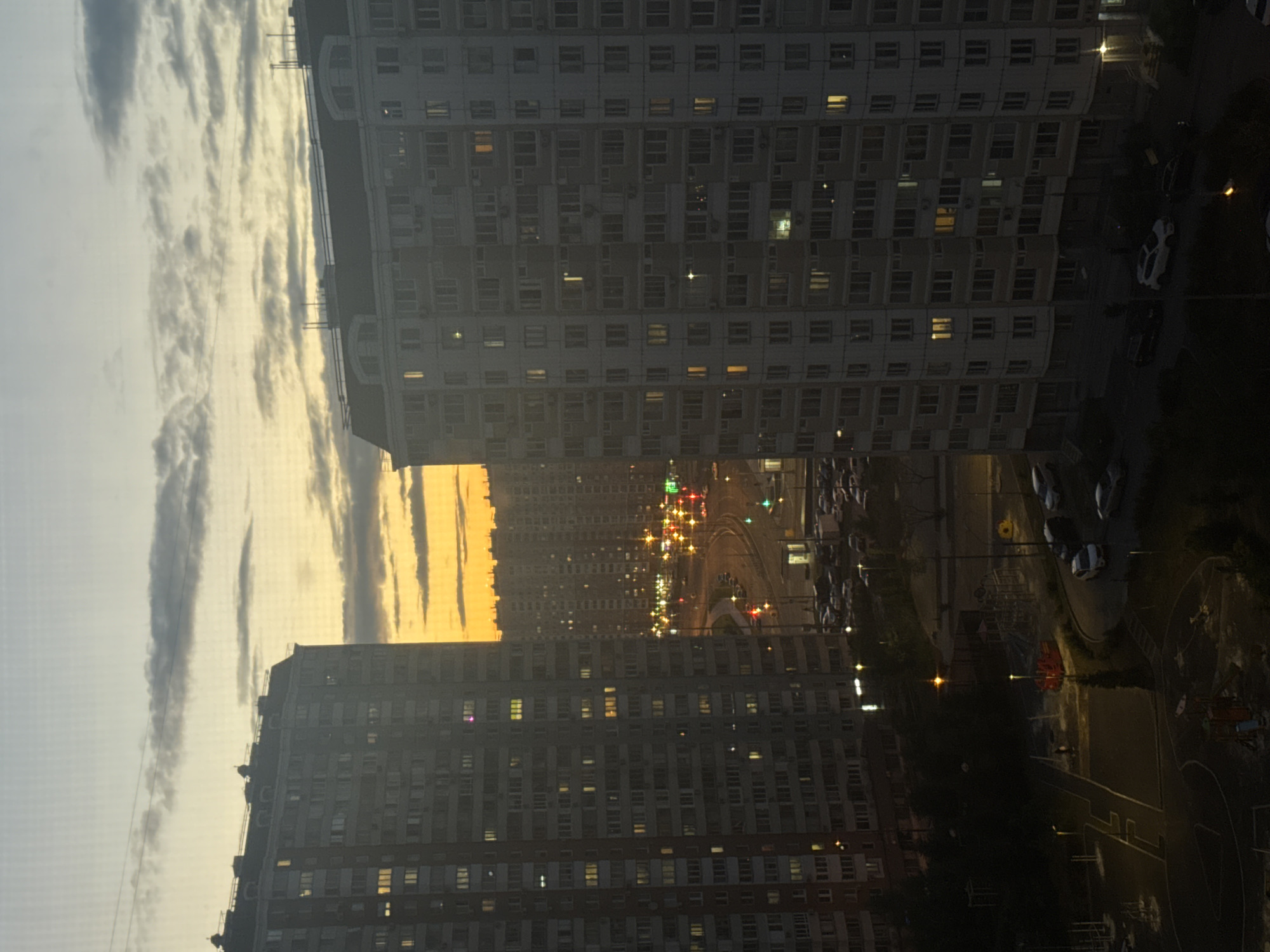 The image depicts a dusk scene in a dense urban area, likely a residential district in Moscow, Russia. Two large, multi-story apartment buildings dominate the foreground, with numerous windows illuminated from within. Between these buildings, a curved road is visible, lined with streetlights and traffic signals displaying red and green lights. Parked cars are scattered along the road and in a nearby parking lot. In the distance, a sprawling cityscape of similar apartment buildings extends towards a horizon illuminated by a bright orange and yellow sunset sky, with scattered clouds. The lower portion of the image reveals a dark courtyard area with playground equipment and more parked cars. The overall impression is one of a typical evening in a large, modern Russian city.