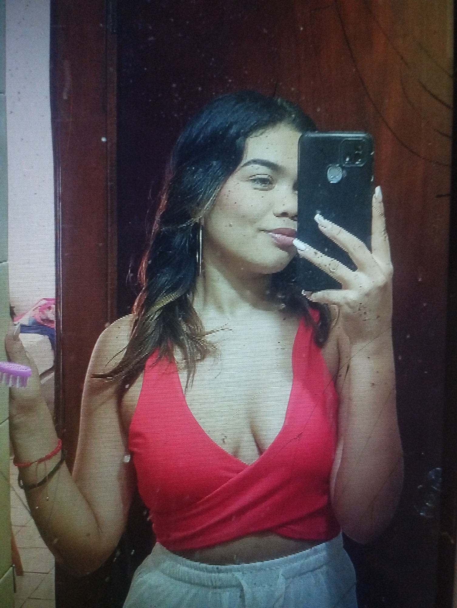 A young woman is taking a selfie in what appears to be a bathroom. She is wearing a red crop top and light-colored sweatpants. She is holding a black smartphone in her right hand, partially obscuring her face. Her left hand is holding a pink hairbrush. The background shows a tiled wall and a dark-colored wooden door. There is no information available in the image to identify the location as Punta de Piedras, Venezuela.
