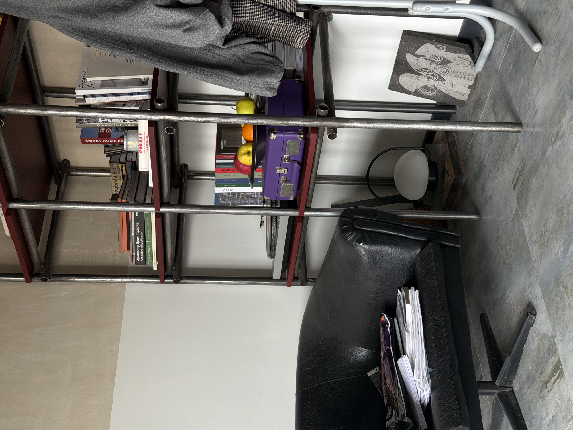 The image shows a close-up of a metal bookshelf with books, fruit, and other objects. The books include titles in English and Russian, such as "French Art," "SMART HOME EX," "COUNTRY CHIC LIVING," "UTOPIA & COLLAPSE," and "Архитектор Джим Торосян" (Architect Jim Torosyan). There is a small purple suitcase, a white lamp, and a stone tile with an image on it. A gray coat or jacket hangs on the upper shelves. A black leather chair sits to the right. The environment suggests an interior space. The image does not provide information about the location being in Yerevan, Armenia, nor does it include details about people, actions, or interactions.