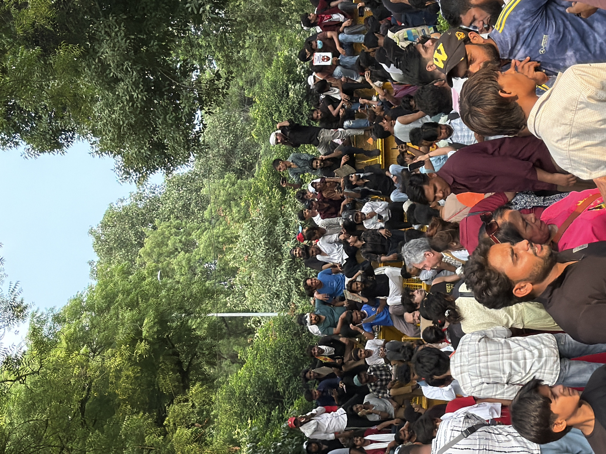 A medium shot captures a crowd of people gathered outdoors, possibly for an event or protest, under a canopy of green trees and a bright sky. The crowd appears to be predominantly male, with many individuals dressed in casual attire. Some people are seated on a raised platform, while others stand or move through the area. The scene has a dynamic feel, with a mix of individuals engaged in conversation, using their phones, or observing their surroundings. The environment suggests a public space, likely in the city of New Delhi, India, given the context provided. The natural elements of trees and sky indicate an outdoor setting.