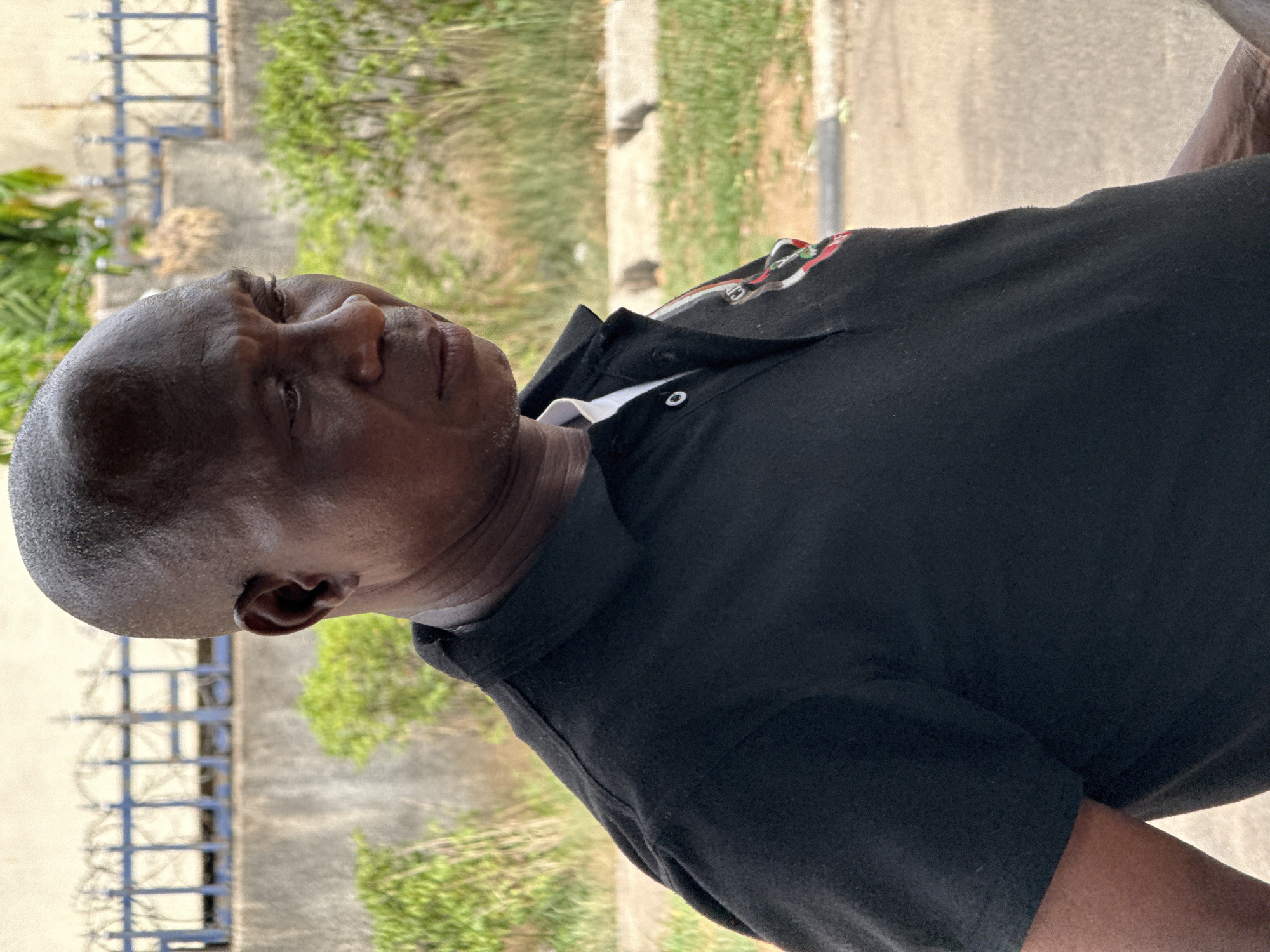 A medium shot captures an older, bald, dark-skinned man in a black polo shirt, looking upwards and to the left. The man is positioned in front of a gated fence with green vegetation behind it, which is on a street in Abuja, Nigeria. The environment suggests an outdoor setting, potentially near a residence or building, as evidenced by the fence and greenery.