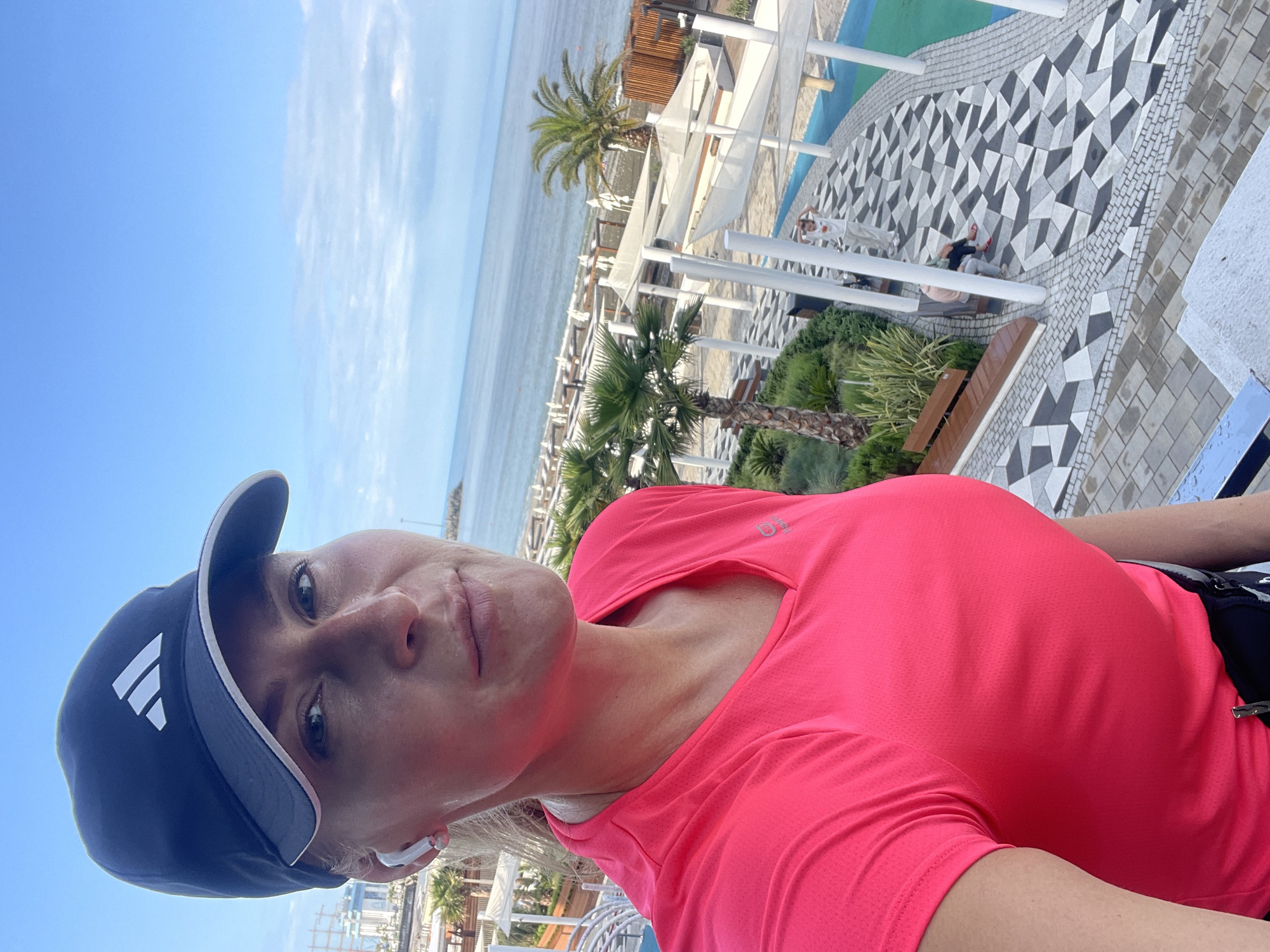 A woman wearing a pink shirt and a blue Adidas baseball cap is seen in a selfie-style shot taken from a high vantage point in what appears to be Sochi, Russia. She's wearing wireless earbuds. Behind her, there's a beach resort area with palm trees and white lounge areas. People can be seen lounging on the ground in the background. The sea and sky are visible in the distance. The ground near the camera features a geometric black and white patterned design.