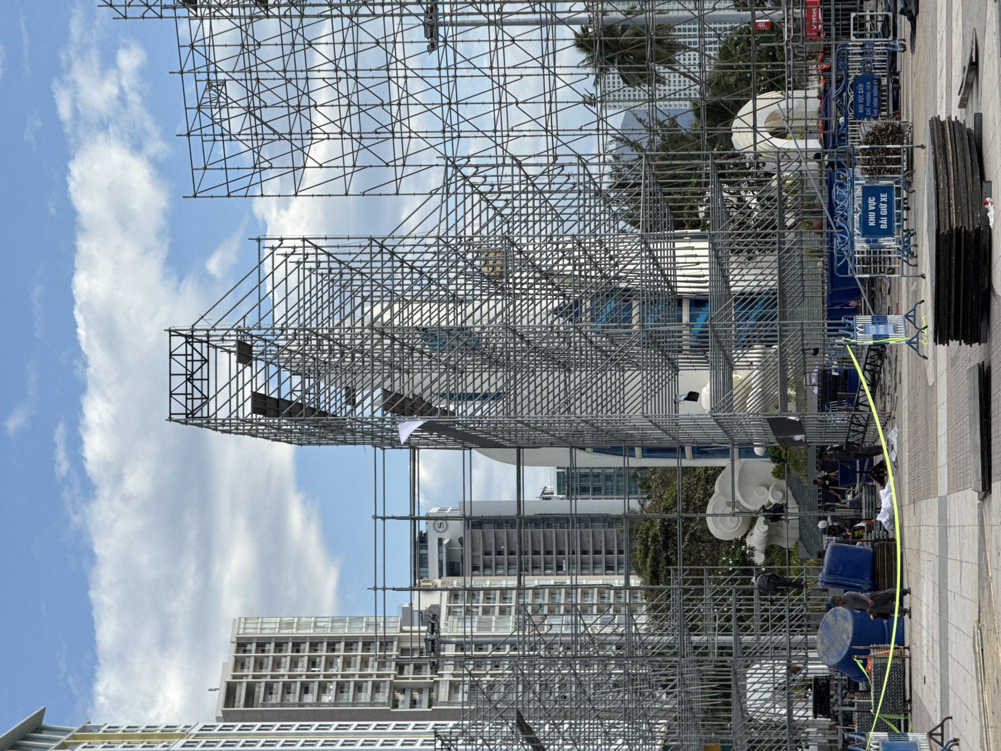 The image depicts a construction site in Nha Trang, Vietnam, characterized by a vast network of scaffolding. Tall modern buildings with multiple floors and windows rise in the background against a sky with scattered white clouds. In the foreground, the scaffolding forms an intricate structure, partially obscuring a white sculptural element that resembles a large cloud or wave. Several large blue plastic containers and stacks of wooden planks are visible on the paved ground. A bright yellow caution tape stretches across the area, indicating restricted access. There are at least five individuals present, some of whom appear to be workers engaged in construction activities. One man in dark clothing is climbing the scaffolding. Another man in similar attire is walking across the paved area. A group of people, some in dark uniforms, are gathered near the blue containers and a fence. The environment suggests an ongoing construction project, possibly for an event or a permanent structure, in an urban setting. Two blue signs with Vietnamese text are posted on metal barriers.