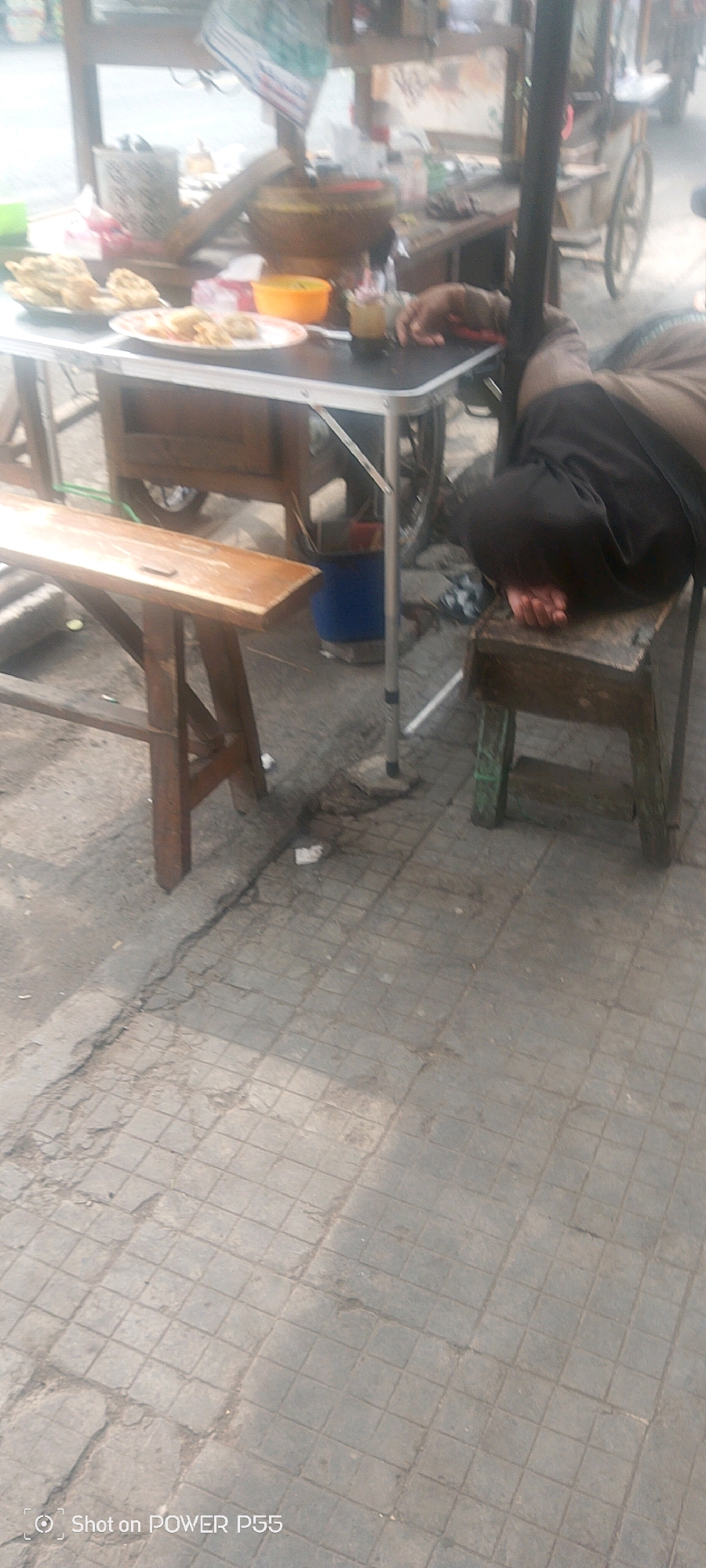 An outdoor scene depicts a food cart with various food items on plates and in containers. A person is seen sleeping on a wooden stool next to the cart. The ground is paved with square tiles, and a wooden bench is visible to the left of the cart. The scene appears to be a street or sidewalk in an urban area.