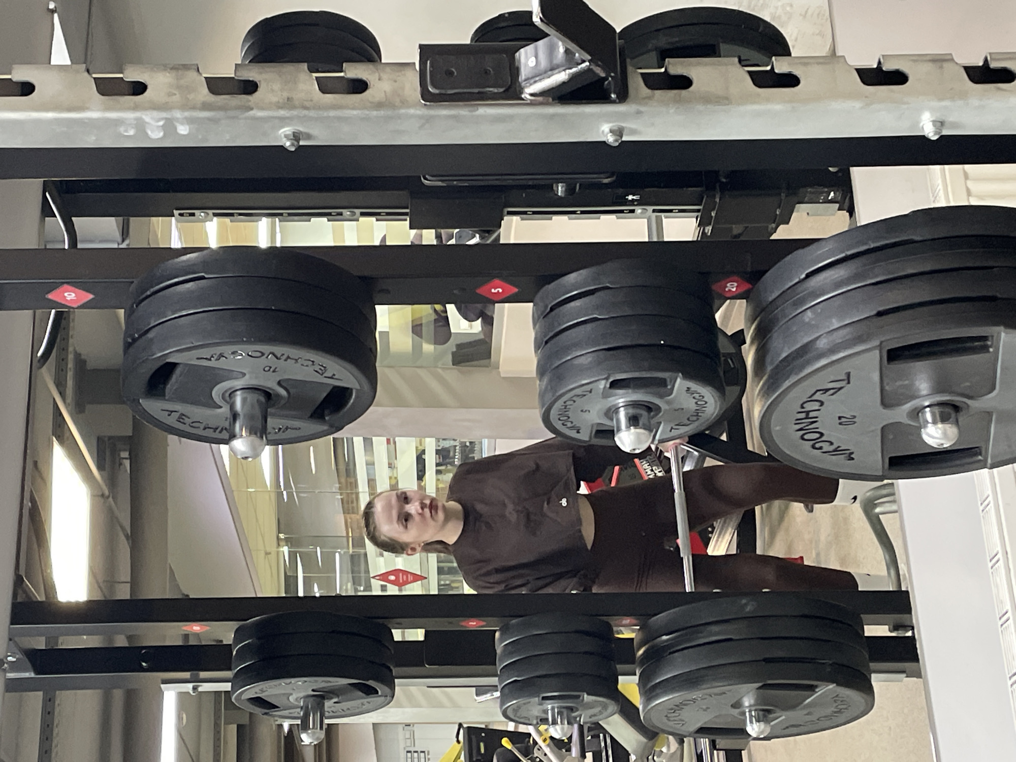 The image depicts an indoor gym setting, likely in Izhevsk, Russia. Dominating the foreground is a weight rack with black weight plates stacked on multiple levels. Each plate is marked with the brand name "TECHNOGYM" and varying weights, such as 10 and 20.
Between the weight racks, a person is visible, wearing a brown workout outfit consisting of a cropped long-sleeve top and leggings. The person appears to be looking towards the camera.
The background suggests a gym environment with other equipment and fixtures, including white vertical panels. The overall lighting is bright and diffused.