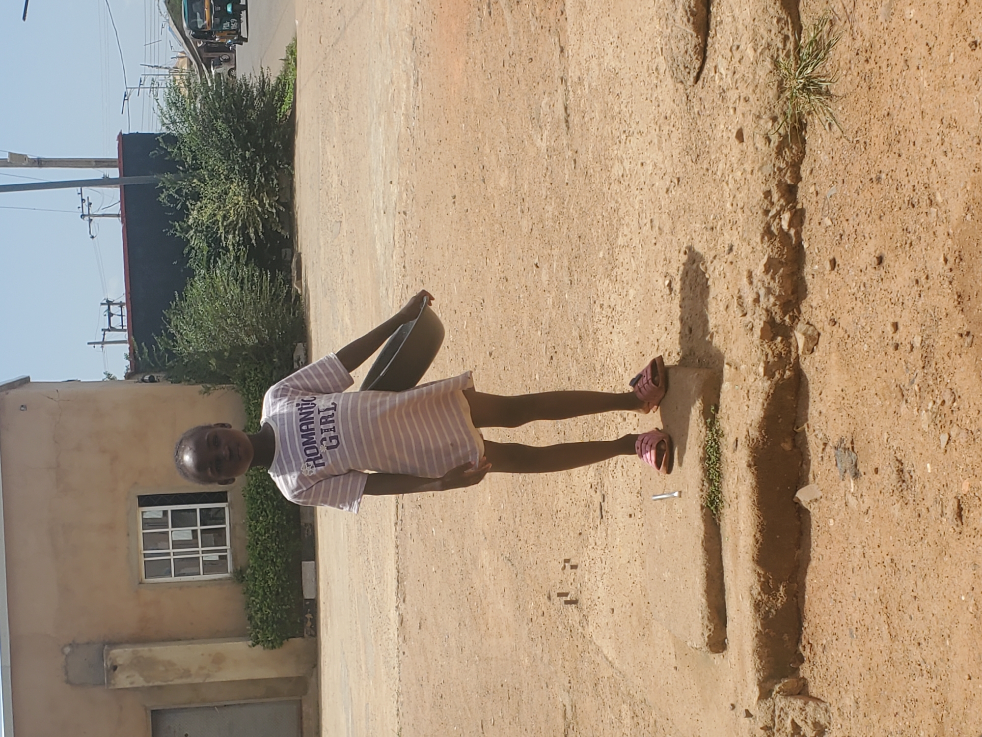 A young girl stands on a dirt road in Jos, Nigeria, holding a bowl. She is wearing a pink and white striped shirt that says "Romantic Girl" and pink sandals. The environment is composed of a light-colored building with a window, green bushes, and a cracked dirt road. A tricycle is visible in the distance.