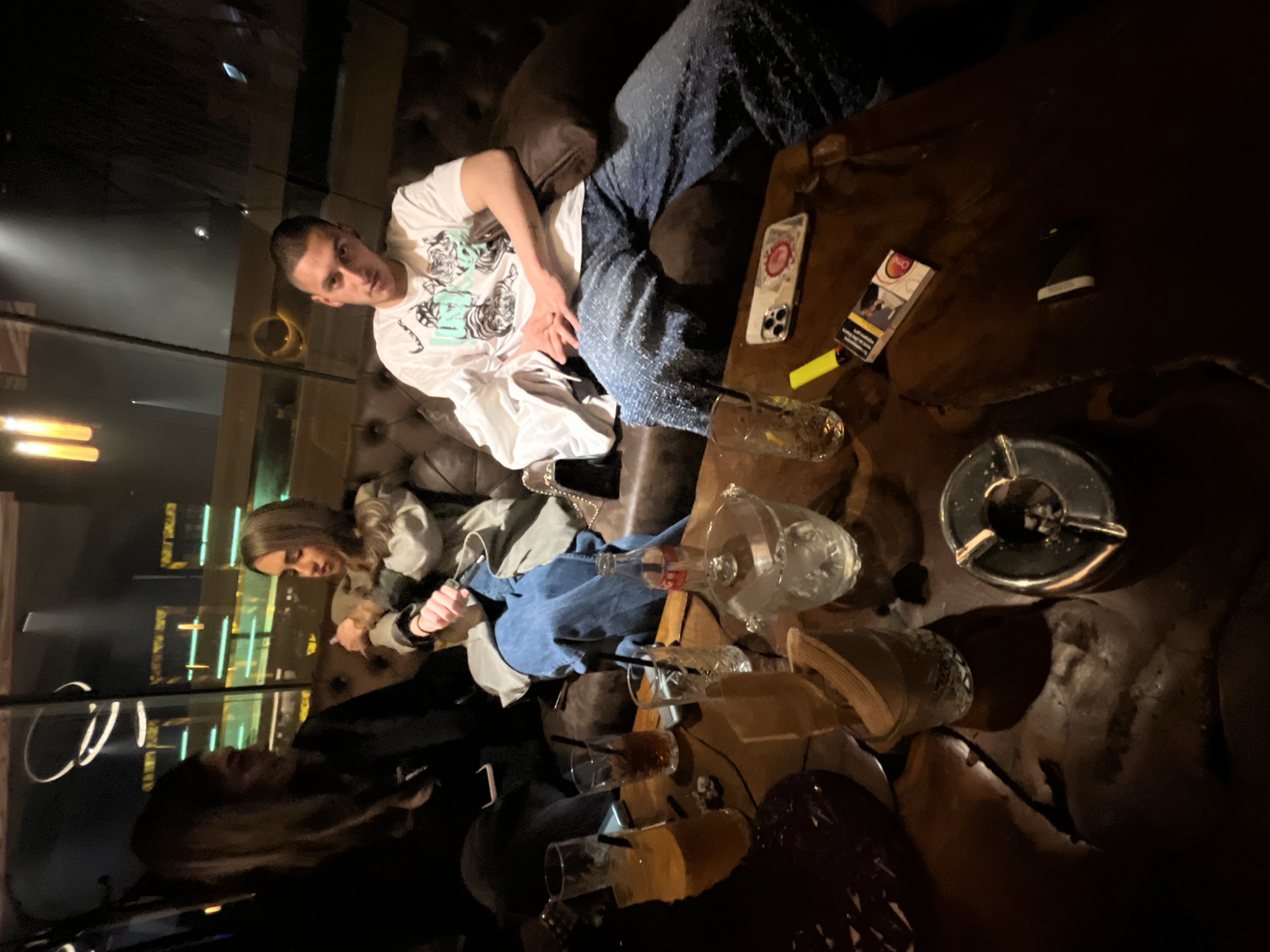 The image shows an interior scene, likely a bar or lounge, with dim lighting. Three people are visible, casually dressed, sitting on a plush brown leather couch. The man on the right is wearing a white t-shirt and blue jeans. A woman is on the left, wearing a grey jacket and jeans. A second woman is in dark clothing. The table in front of them is covered with multiple empty glasses, an ashtray, a pack of cigarettes, a lighter, and a smartphone. The background features a window or wall with illuminated vertical lines, suggesting an urban setting. There is no information to confirm the location in Burgas, Bulgaria.