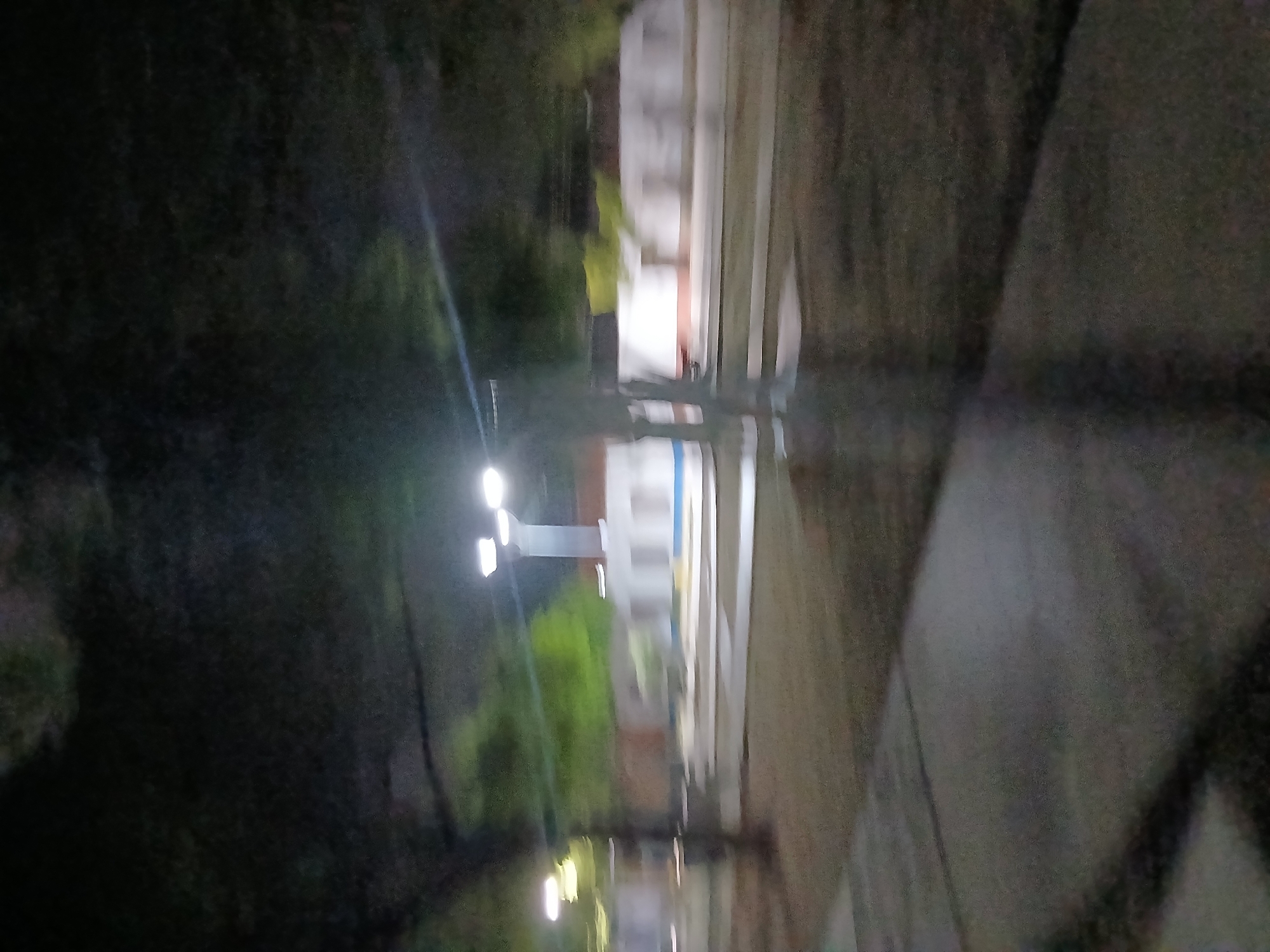 A blurry, nighttime exterior shot shows a pathway in front of a low-rise building with white walls and a red roof. A three-headed lamppost is visible on the right side of the frame, illuminating the area. Trees and foliage are present in the background, and the ground appears to be paved or covered in gravel. The scene is indistinct due to motion blur.
