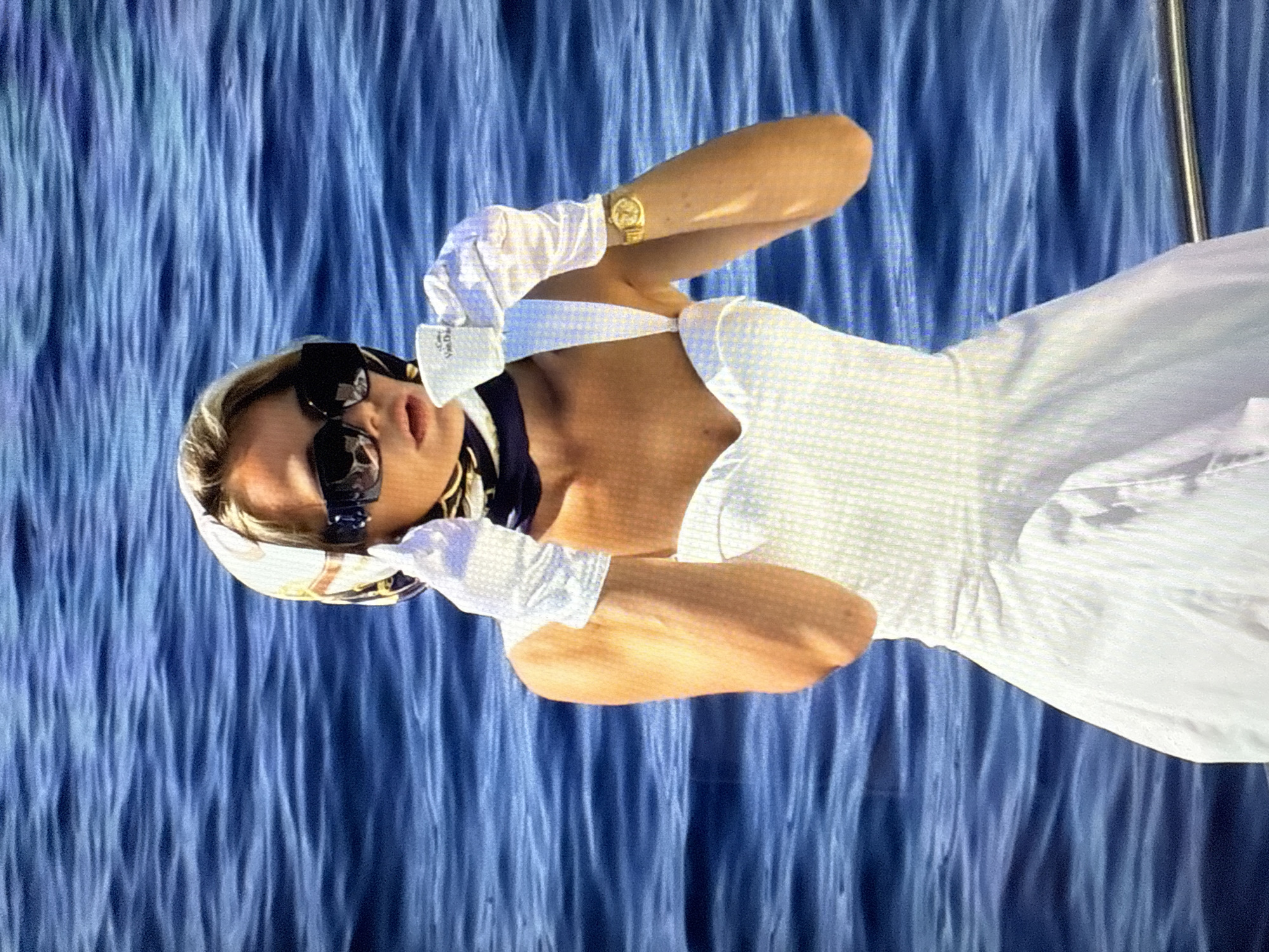 A blonde woman wearing a white dress, white gloves, sunglasses, and a headscarf stands on a boat on the water. She is holding a small white cup and appears to be drinking from it. The water is blue and choppy. The location is not specified as Găvănoasa, Moldova, as there is no visual information to support this claim.