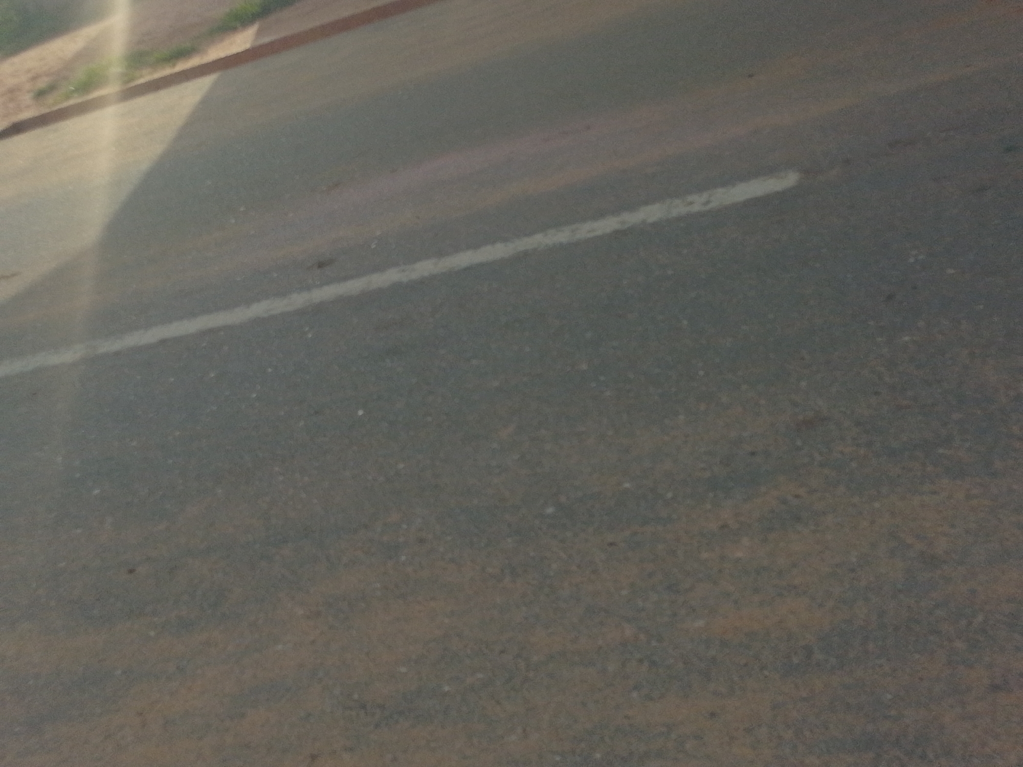 The image shows a close-up, angled view of an asphalt road with a white line marking the edge. The road surface is dark gray and appears to be slightly dusty or worn, with small flecks of lighter colored material visible. A thin, white line runs diagonally across the lower half of the frame, indicating a lane or road boundary. To the upper left, a strip of reddish-brown earth or brickwork lines the edge of the road, with a small amount of dry, light green vegetation visible on top of it. A bright, vertical light flare emanates from the upper left corner, casting a shadow across the road surface, obscuring details in that area. The lighting suggests it is daytime. The scene appears to be an outdoor, urban or suburban environment in Tempure, Nigeria, characterized by a typical road infrastructure. There are no people or vehicles visible in the frame.