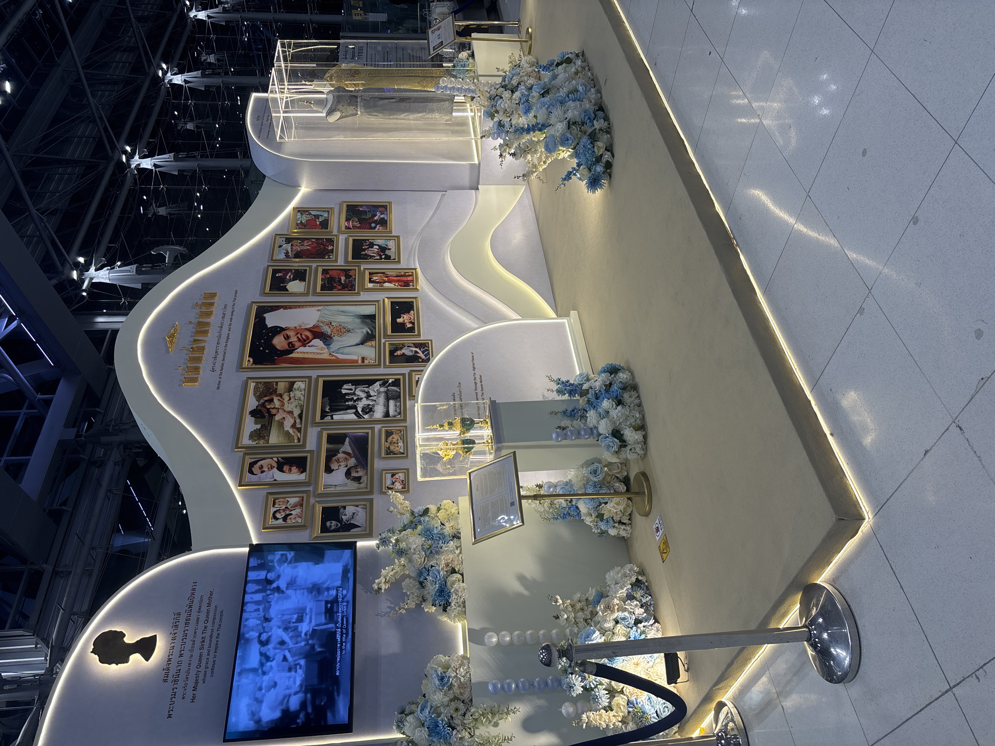 This image captures an exhibition likely held in a public space within Nong Prue Subdistrict, Thailand, given the Thai language text present. The exhibition features a large display dedicated to a prominent Thai figure, identified by the text as "Her Majesty Queen Sirikit, The Queen Mother."
The central element of the display is a curved, illuminated wall adorned with numerous framed photographs. These photographs appear to depict the Queen Mother throughout her life, in various settings and with family members. A large screen on the left side of the display plays a video, also likely related to the Queen Mother.
Flanking the central display are elegant floral arrangements in shades of blue and white, enhancing the celebratory and respectful atmosphere. To the right, glass display cases house what appear to be ornate artifacts, possibly royal regalia or significant personal items. A mannequin is visible within a display case, wearing a traditional Thai garment.
The exhibition is set up on a raised platform covered with light-colored carpeting, which is outlined by subtle LED lighting. Stanchions with velvet ropes are placed in the foreground, indicating a designated viewing area and guiding visitors. The background suggests an indoor environment, possibly an airport terminal or a large exhibition hall, with high ceilings and industrial lighting structures visible. The overall impression is one of a well-curated and visually appealing tribute.