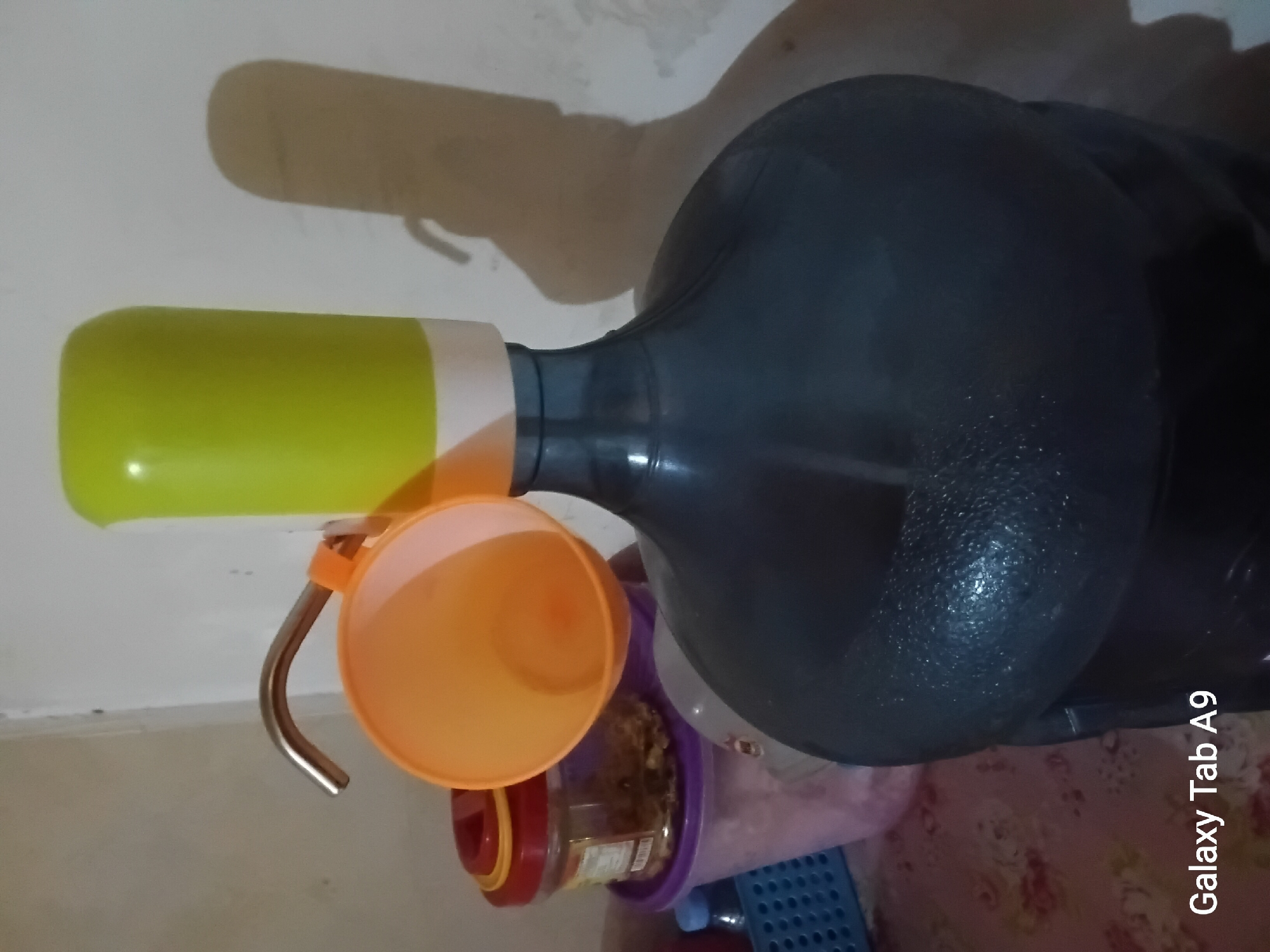 A gallon water dispenser with a green plastic top and a metal spout is the central subject. An orange plastic cup is attached to the spout. In the background, there are several storage containers, including one with a red lid and another with a purple lid. A blue basket is visible to the left. The scene appears to be indoors, possibly in a home or small shop, with a light-colored wall and a patterned tablecloth. The lighting creates a shadow of the dispenser on the wall. The timestamp "Galaxy Tab A9" is present in the lower-left corner. The image quality is moderate. There are no people visible.