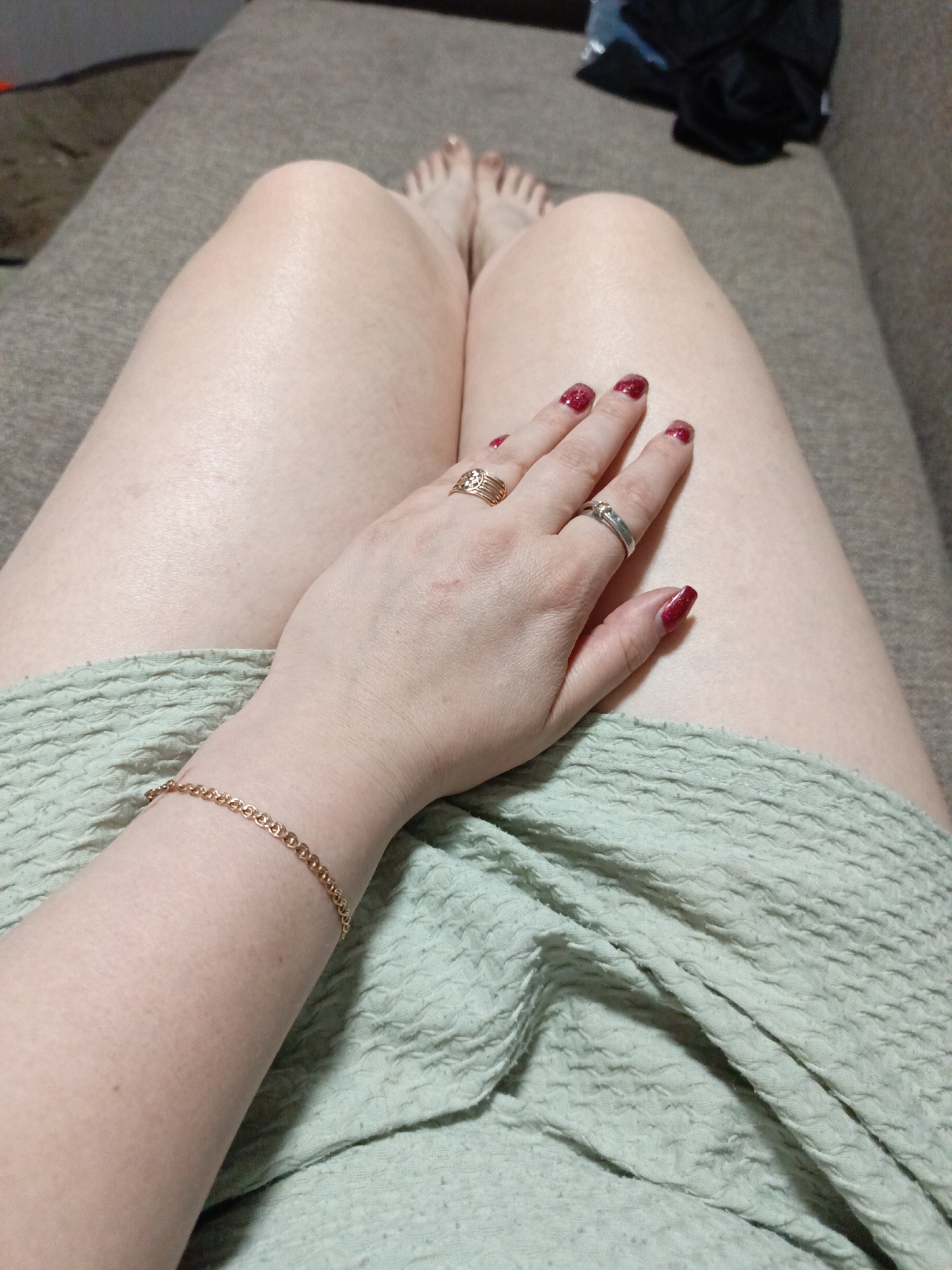 A woman is sitting on a beige couch. She is wearing a gold bracelet and two rings. Her nails are painted red. A green textured blanket covers her lap. In the background, there is a black bag. There is no evidence suggesting the image was taken in or near Марковский, Russia.