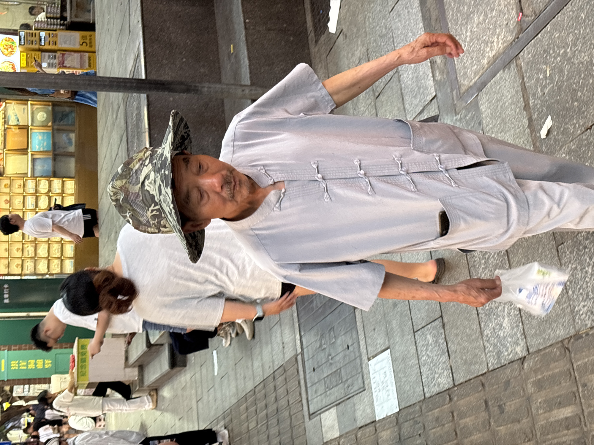 The image depicts a street scene in what appears to be Yuzhong District, China. In the foreground, an older man is the central focus. He is wearing a grey, traditional-style tunic and pants, and a camouflage bucket hat. He has a mustache and is holding a plastic bag in his right hand, with his left arm extended upwards.
Around him are other people. One woman is wearing a white t-shirt and dark pants; a ponytail is visible. Several other individuals are visible in the background, mostly out of focus. There's a shop or building in the background with numerous small boxes or items displayed. The ground consists of patterned paving stones.