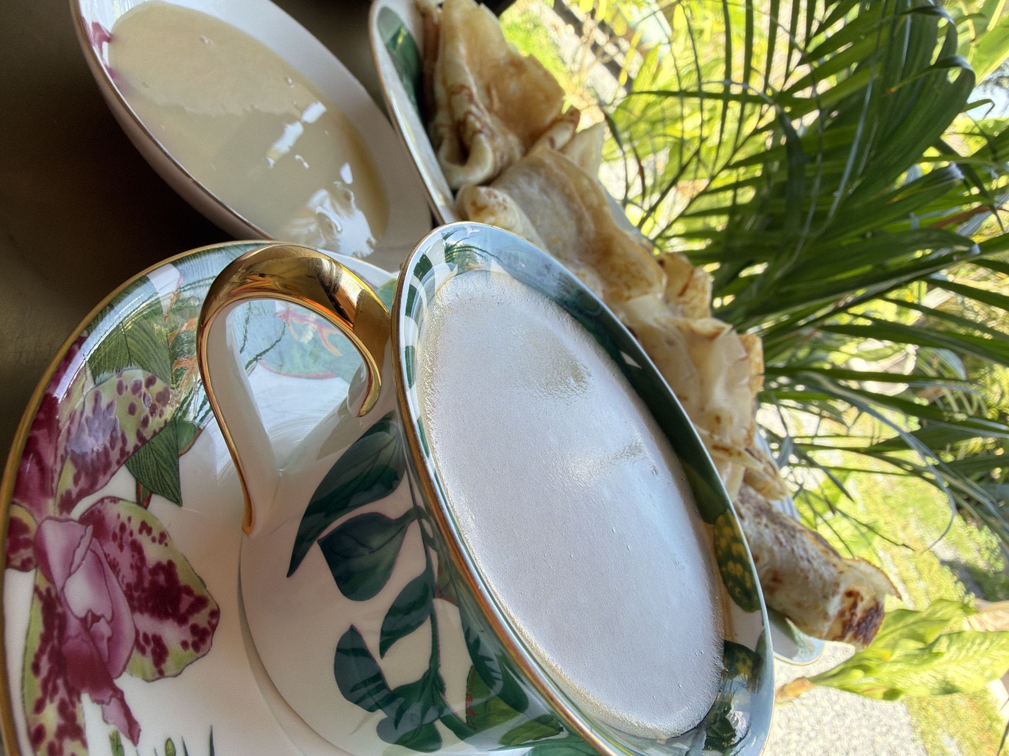 A close-up, eye-level shot shows a fancy tea set and crepes on a table, likely outdoors in Ko Samui District, Thailand, as indicated by the lush green palm fronds and other foliage in the background. A white teacup with a gold rim and a floral pattern of green leaves and pink orchids sits on a matching saucer, filled with a frothy, light-colored beverage. To the left, a white oval dish holds a pale yellow, creamy liquid, possibly condensed milk or a sweet sauce. Behind the teacup and to the left is a plate piled with golden-brown crepes, some rolled and others folded. The lighting suggests a bright, sunny day, with sunlight filtering through the tropical greenery. No people are visible in the image.