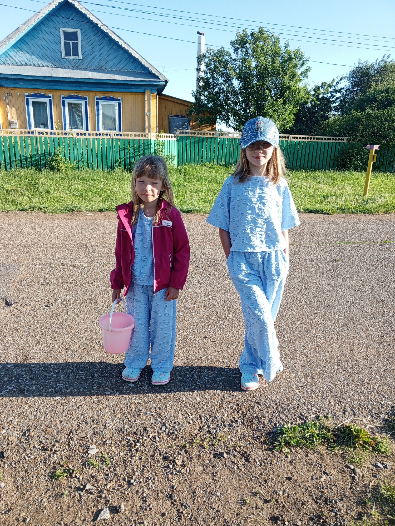 In Buraly, Russia, two young girls stand on a gravel road in front of a yellow house with a blue roof and a green fence. The girl on the left, wearing a pink jacket over a light blue outfit and holding a pink bucket, has blonde hair and is looking towards the camera. The girl on the right, sporting a blue baseball cap and a matching blue patterned outfit, also faces the camera with her hands in her pockets. The environment is rural, with grass and trees visible behind the fence. The sky is clear and blue, indicating a sunny day.