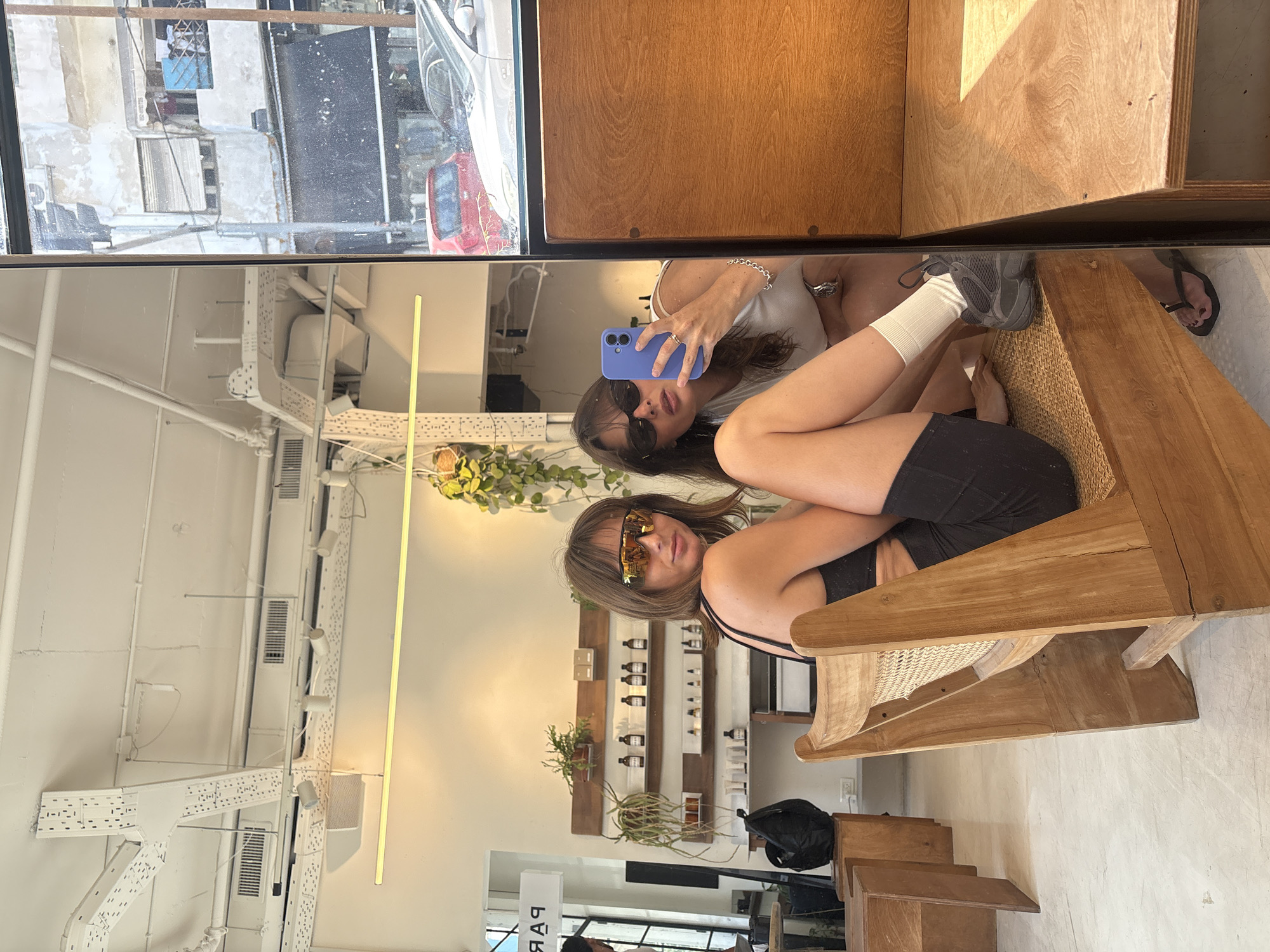 Two young women are seated in chairs within a brightly lit, minimalist establishment, likely a cafe or boutique, in Tel Aviv, Israel. The woman on the left, wearing large, reflective sunglasses and a black tank top, is seated in a wooden chair with a woven seat, looking towards the camera. The woman on the right, visible in the mirror's reflection, holds a blue smartphone in her right hand, taking a selfie. She wears sunglasses and a white top, with white socks and athletic shoes. The environment features exposed ceiling conduits, track lighting, and shelves displaying bottles. A sign with reversed lettering, reading "PAR...", is visible on the left. The reflection in the mirror shows a street scene with buildings and parked cars, characteristic of Tel Aviv.