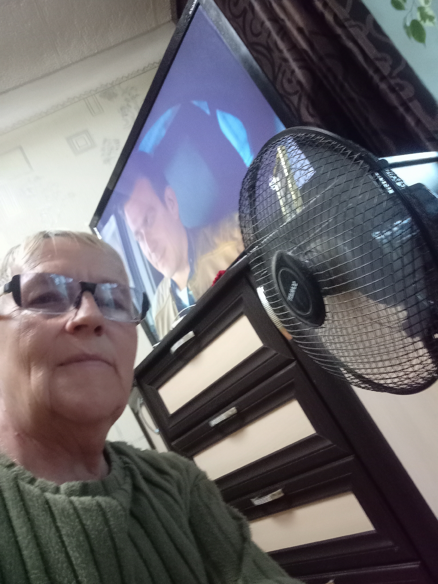 An indoor scene captured from a low-angle perspective shows a person with short blonde hair and glasses in the foreground, wearing a green ribbed sweater. Behind the person, a television displays an image of a man in a yellow jacket. To the right of the television, a black oscillating fan is partially visible, with its grille and motor housing in focus. A dark wooden dresser with beige drawers is situated in front of the television, below the fan. The background shows a textured wall with a patterned curtain. The overall environment suggests a residential interior.