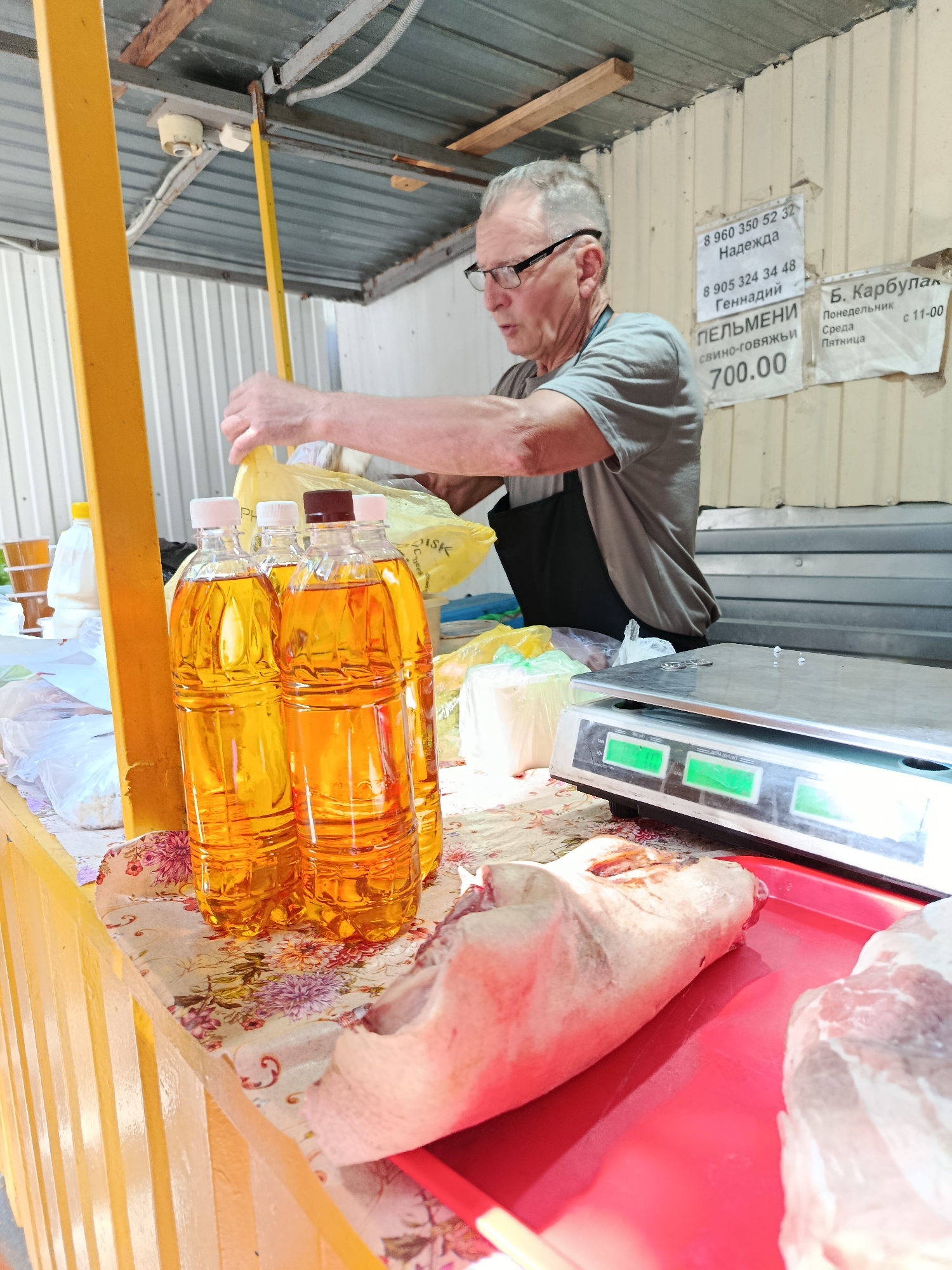 A man with gray hair and glasses, wearing a gray t-shirt and a dark apron, stands behind a market stall in Saratov, Russia. He is handling yellow plastic bags filled with goods. In the foreground, on a table covered with a floral cloth, are several plastic bottles filled with a golden liquid, possibly cooking oil. Next to the bottles are more plastic bags. To the right of the bottles is a digital scale. In front of the scale, on a red tray, are pieces of raw meat, one appearing to be a pig's trotter. Behind the man, a metal roof covers the stall, and a white corrugated wall is visible. Signs with Russian text are affixed to the wall. The overall scene depicts a typical outdoor market setting.